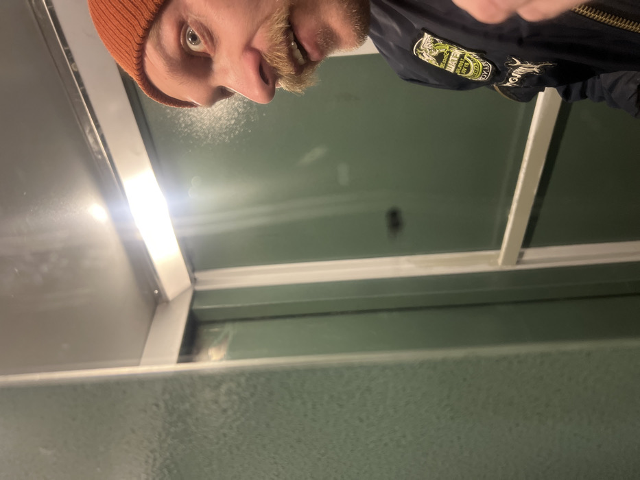 The image is a low-angle shot, likely a selfie, featuring a person with a mustache, wearing an orange beanie and a black jacket with patches. The person's face is close to the camera, showing a slightly grinning expression. The background consists of green wall panels separated by white frames, suggesting an interior space, possibly a public restroom. A bright light fixture is visible near the top-left corner of the frame. The image has a somewhat distorted perspective due to the angle.
