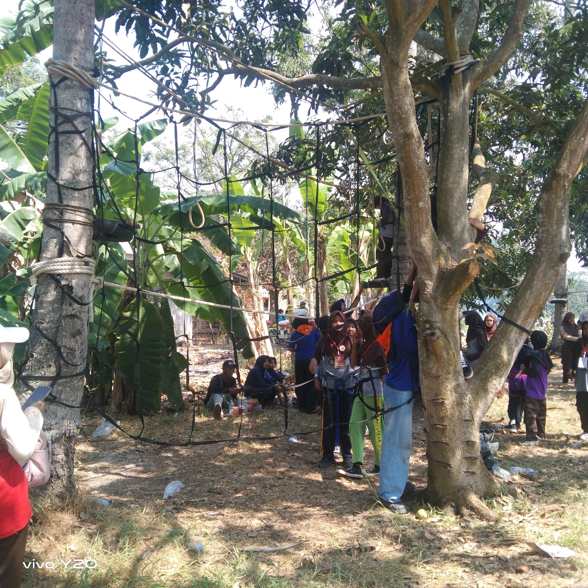 In Karawang, Indonesia, a group of individuals, many of whom appear to be students wearing casual clothing and some in vests and harnesses, are gathered in an outdoor area that seems to be part of an adventure or team-building activity. The environment consists of several trees, including a large one with multiple branches prominently featured in the foreground, and abundant vegetation, such as banana plants. The trees are rigged with ropes and netting, suggesting an obstacle course or climbing setup. Some people are actively engaged with the ropes, either climbing or preparing to climb, while others are observing or resting on the ground. Several individuals are wearing headscarves, indicating a cultural context. The ground is covered with dry leaves and grass, and scattered plastic bottles are visible, suggesting a somewhat informal setting. The overall scene depicts a recreational or educational event taking place in a natural, outdoor environment.