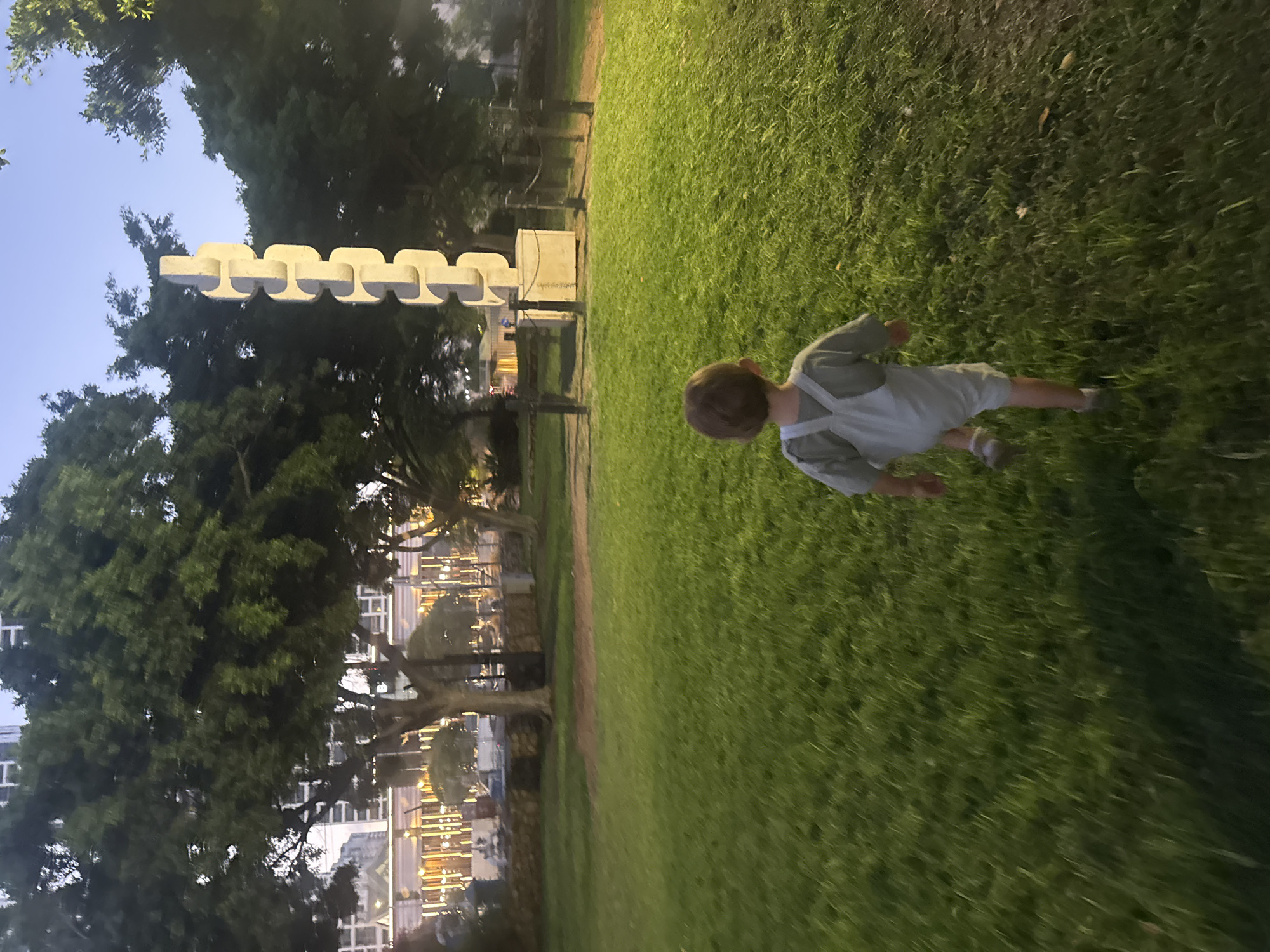 In the city of Bat Yam, Israel, a young child wearing light-colored overalls runs across a grassy field at dusk. The child is facing away from the camera, heading towards a large, abstract concrete sculpture that resembles a stack of interlocking links. The sculpture stands on a concrete base, surrounded by a low fence. Behind the sculpture and the child, tall trees line the edge of the park. In the background, lights from buildings and streetlights illuminate the scene. The sky is a dusky blue, indicating the transition from day to night. The overall impression is of a peaceful evening scene in a public park.