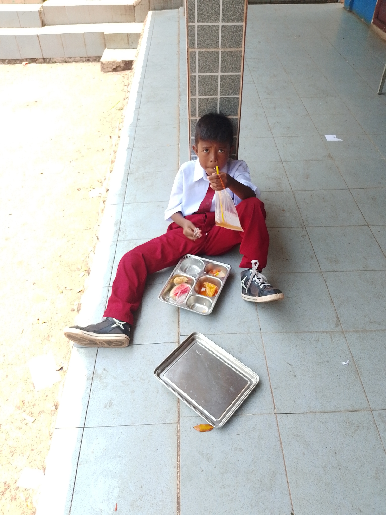 An outdoor shot shows a young boy in a white collared shirt and red pants sitting on a tiled walkway next to a wall. He is holding a plastic bag with a yellow straw and appears to be drinking from it. In front of him are two metal trays. The tray closer to him contains food. A second, empty tray is positioned to his right. The walkway is made of light-colored square tiles and is partially in shadow. To the left of the boy, there is a sandy area with some foliage. The wall behind him has a pattern of dark grey squares. The scene appears to be in a school or public area in Batam, Indonesia.