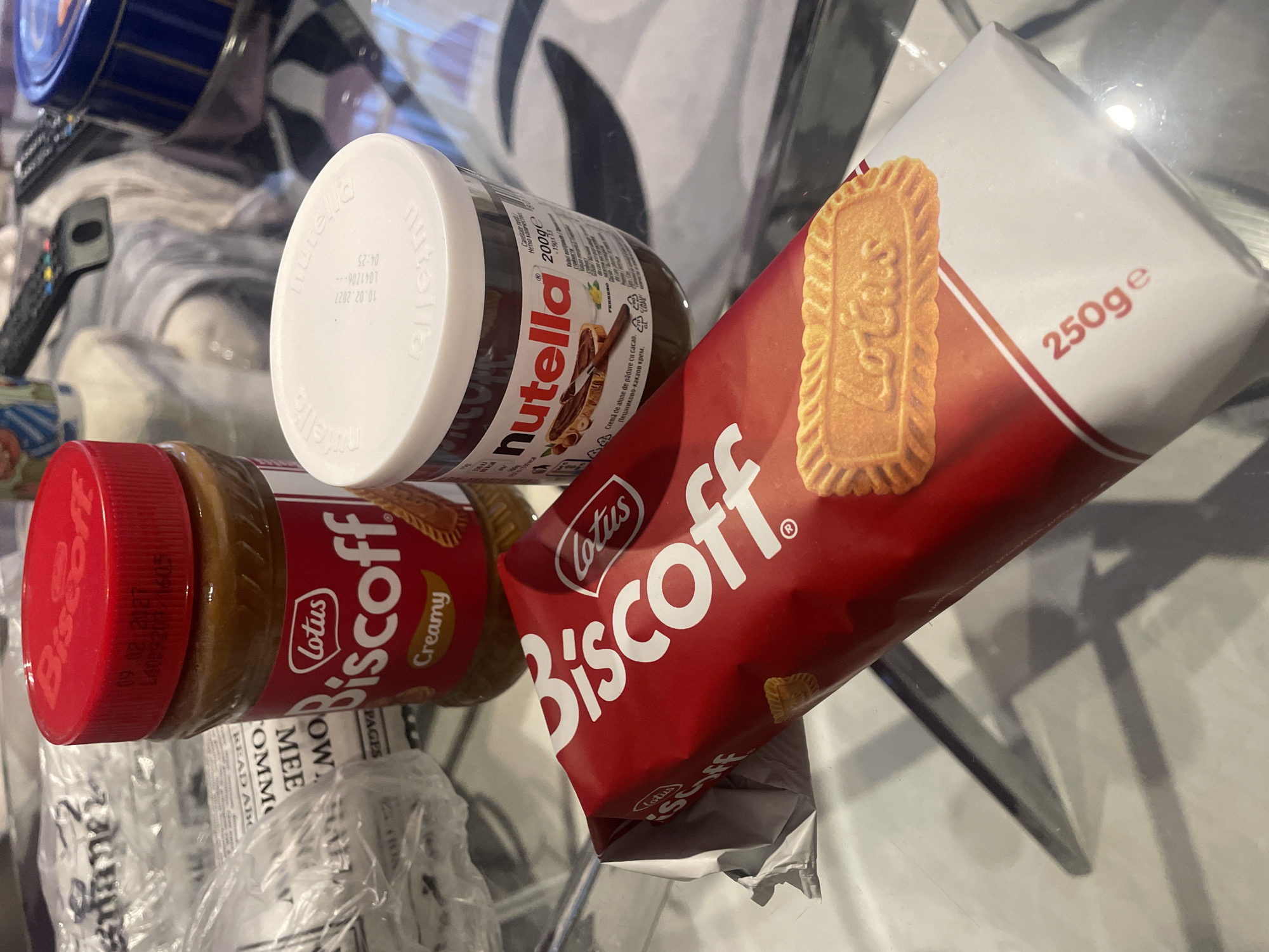The image shows a collection of food items on a glass surface. There is a jar of Nutella, a jar of Lotus Biscoff spread, and a package of Lotus Biscoff cookies. The glass surface reflects some of the objects above it. The OCR of the image shows the date "10.02.2027" on the lid of the Nutella jar and "09.02.2027" on the lid of the Biscoff spread. The package of Biscoff cookies is labeled as 250g. No people are visible. The context of the location in Sofia, Bulgaria, is based on the OCR text "Пешниково-какаов крем." which is the Bulgarian phrase for hazelnut cocoa cream.