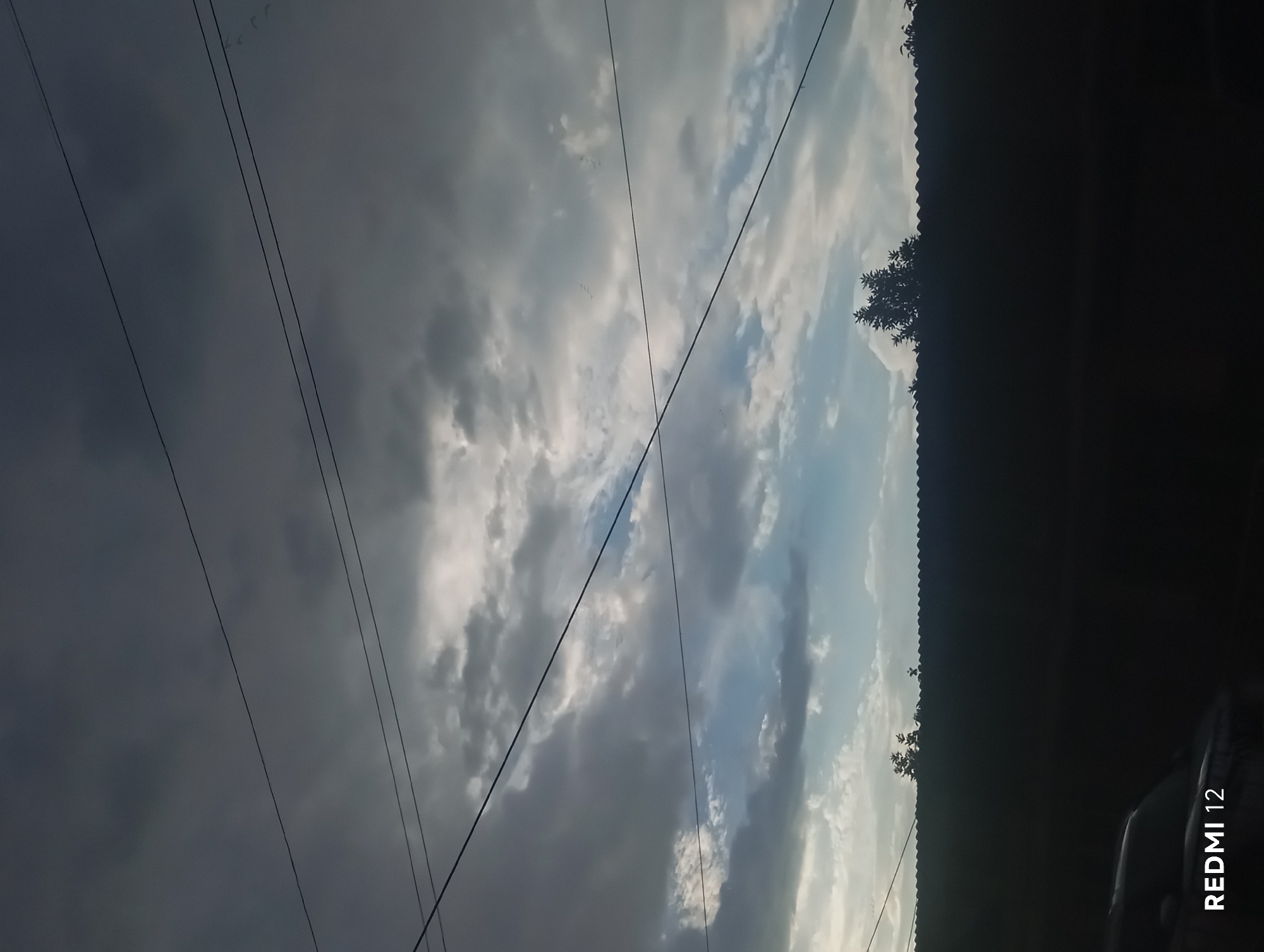 The image depicts a dramatic sky with a mix of dark, brooding clouds and brighter, sunlit patches. The sky is filled with textured clouds, some appearing heavy and overcast, while others are lighter and more wispy, allowing glimpses of blue sky. The overall impression is one of dynamic weather, possibly before or after a storm.
Several thin, dark lines, likely power lines, run diagonally across the frame, intersecting the sky. In the upper left quadrant, a scattering of small, faint specks might be birds in flight, adding a subtle sense of scale and movement to the vast sky.
The lower portion of the image is dominated by a dark, silhouetted area. Along the very bottom right, part of a dark vehicle, possibly a car, is visible. The edge of what appears to be a corrugated roof or structure runs horizontally across the middle ground, also in silhouette against the sky. A few silhouetted trees with sparse foliage can be seen along this edge.
The lighting suggests it is either dawn or dusk, with the sun low on the horizon, casting dramatic shadows and illuminating the clouds in a textured way. The time of day is further indicated by the "REDMI 12" watermark in the lower right corner, a typical stamp from a smartphone camera that often includes the time or date. The overall mood is somewhat somber due to the dark clouds and silhouettes, but also awe-inspiring due to the grandeur of the sky.