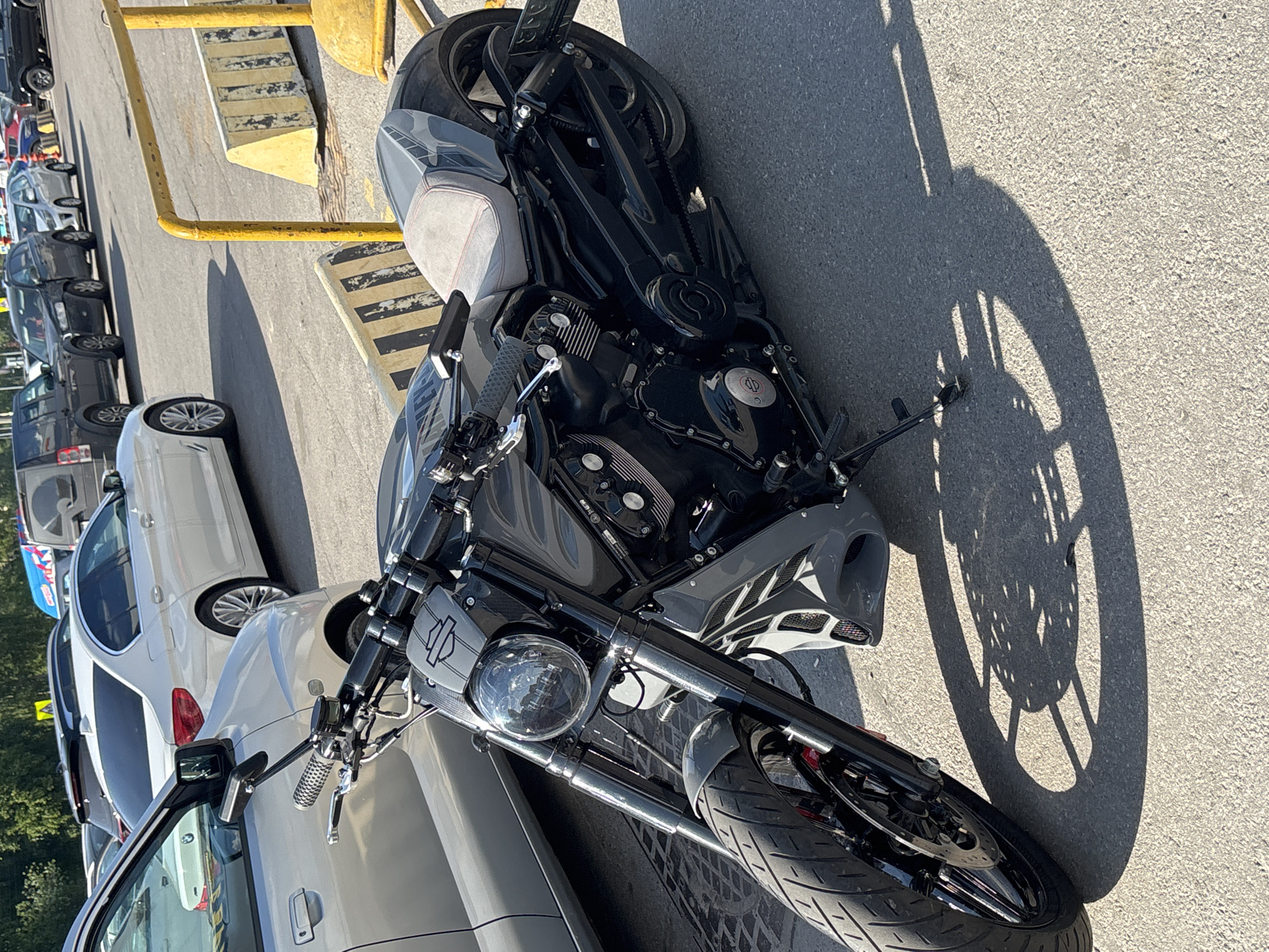 A dark gray Harley-Davidson motorcycle is prominently featured in the foreground of this outdoor, sunlit image. The motorcycle is positioned diagonally, with its front wheel and headlight facing towards the bottom right of the frame. Its engine, exhaust pipes, and black wheels are clearly visible. A gray seat with subtle red stitching is also noticeable. The "HARLEY DAVIDSON" logo is visible on the front part of the motorcycle, above the round headlight.
The motorcycle casts a long, distinct shadow to its right on the asphalt ground, highlighting the spokes of its wheels. Behind the motorcycle, a line of various parked cars extends into the distance, suggesting a parking lot or street setting. Several of these cars are dark-colored, while a white sedan with distinctive wheels is visible in the mid-ground. Bright yellow metal barriers or poles run along the left side of the frame, separating the parked vehicles from what appears to be a curb or walkway.
The scene is brightly lit by direct sunlight, indicating a clear, sunny day, likely during the morning or afternoon given the strong, defined shadows. There are no people visible in the image. The overall impression is one of a stationary motorcycle and other vehicles parked in an urban or semi-urban outdoor environment.
