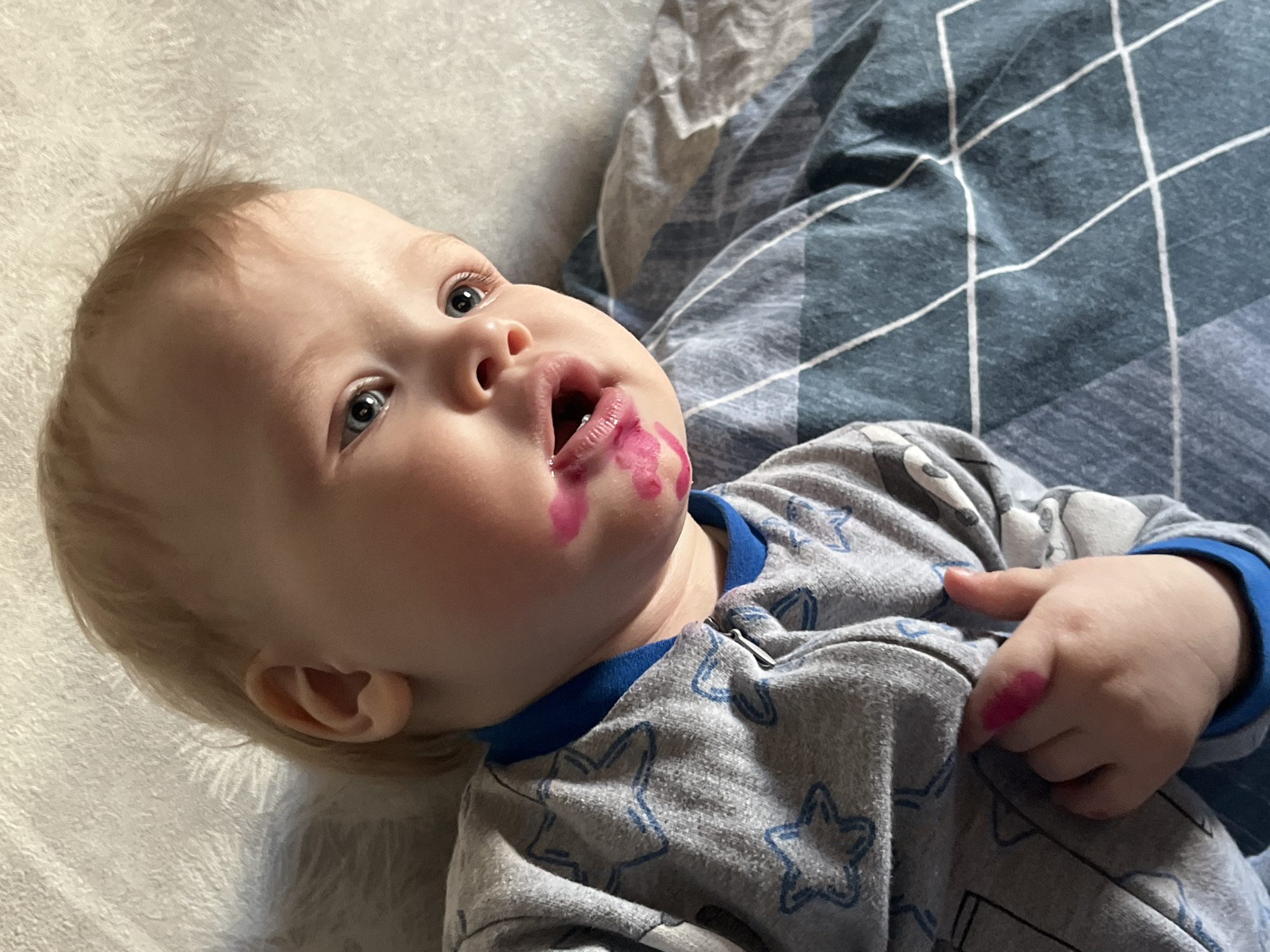 A baby lies on their back on a bed. The baby has fair skin, blond hair, and blue eyes. There are pink stains around the baby's mouth and on their right hand. The baby is wearing a gray pajama set with blue trim and a star pattern. The bed has two different types of bedding: a white, textured blanket on the left and a blue-gray plaid blanket on the right. There is no indication of Chaikovsky, Russia.
