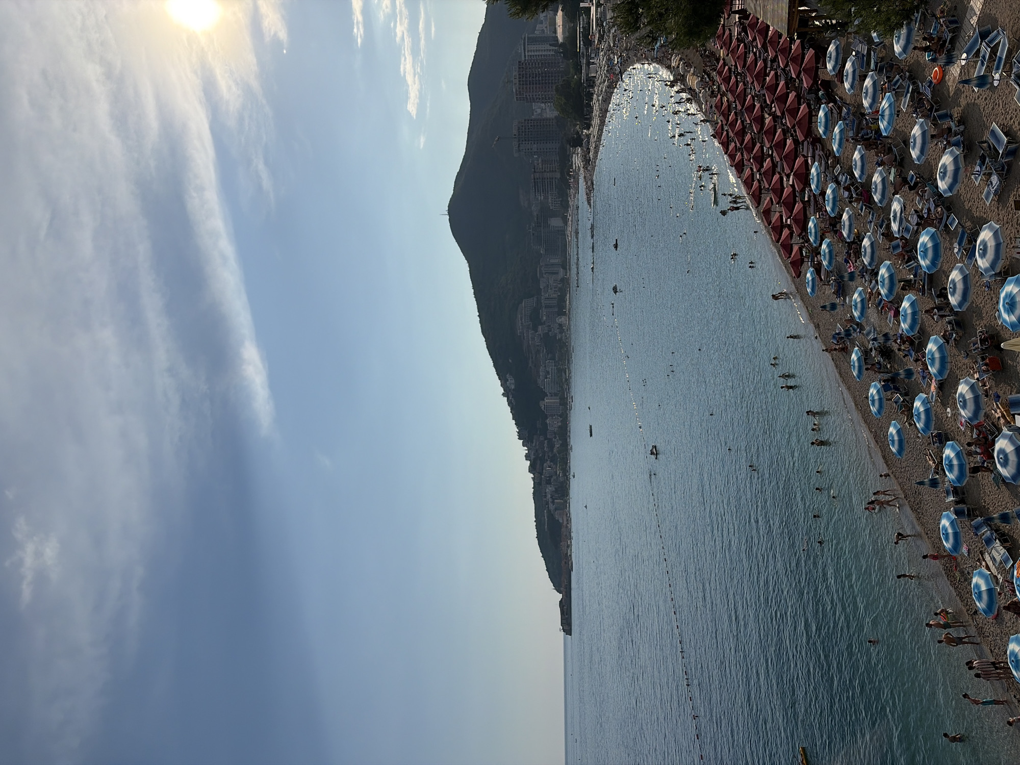 This is a high-angle, eye-level shot of a crowded beach in Budva, Montenegro, with the Adriatic Sea on the left and a town nestled on a hillside in the background, under a partially cloudy sky. The sun is visible in the upper right corner, casting a warm glow. The beach is densely packed with rows of red and blue-and-white striped umbrellas, with lounge chairs beneath many of them. People are scattered across the sand and in the shallow water, some swimming, some sunbathing, and others walking along the shore. A rope barrier separates the swimming area from a deeper part of the sea, where a few small boats and watercraft are visible. The city of Budva extends along the coastline, with buildings of varying heights dotting the green, mountainous terrain that rises behind it.