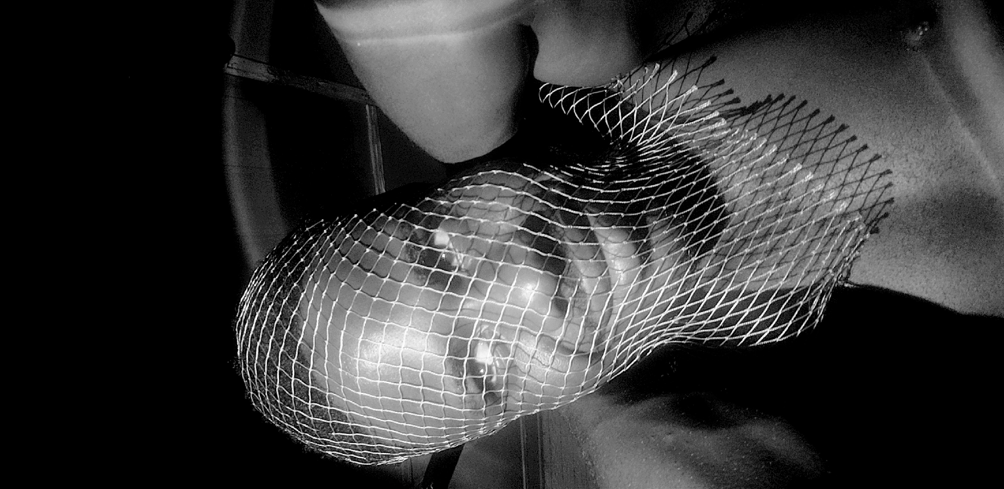 In this black and white, high-contrast, low-light photograph, a person's face is partially obscured by a white mesh net covering their head. The net creates a grid pattern over the individual's features, with some areas of the face, such as the eyes and nose, more visible than others. The lighting emphasizes the texture of the net and the contours of the face beneath it. The background is dark and indistinct, suggesting an indoor setting. There are no discernible landmarks that place the image specifically in Narayi, Nigeria, or any other city. The subject's gender and specific age are not clearly ascertainable due to the netting and lighting. The image appears to be a close-up portrait, focusing on the interplay of light, shadow, and the texture of the netting against the skin.