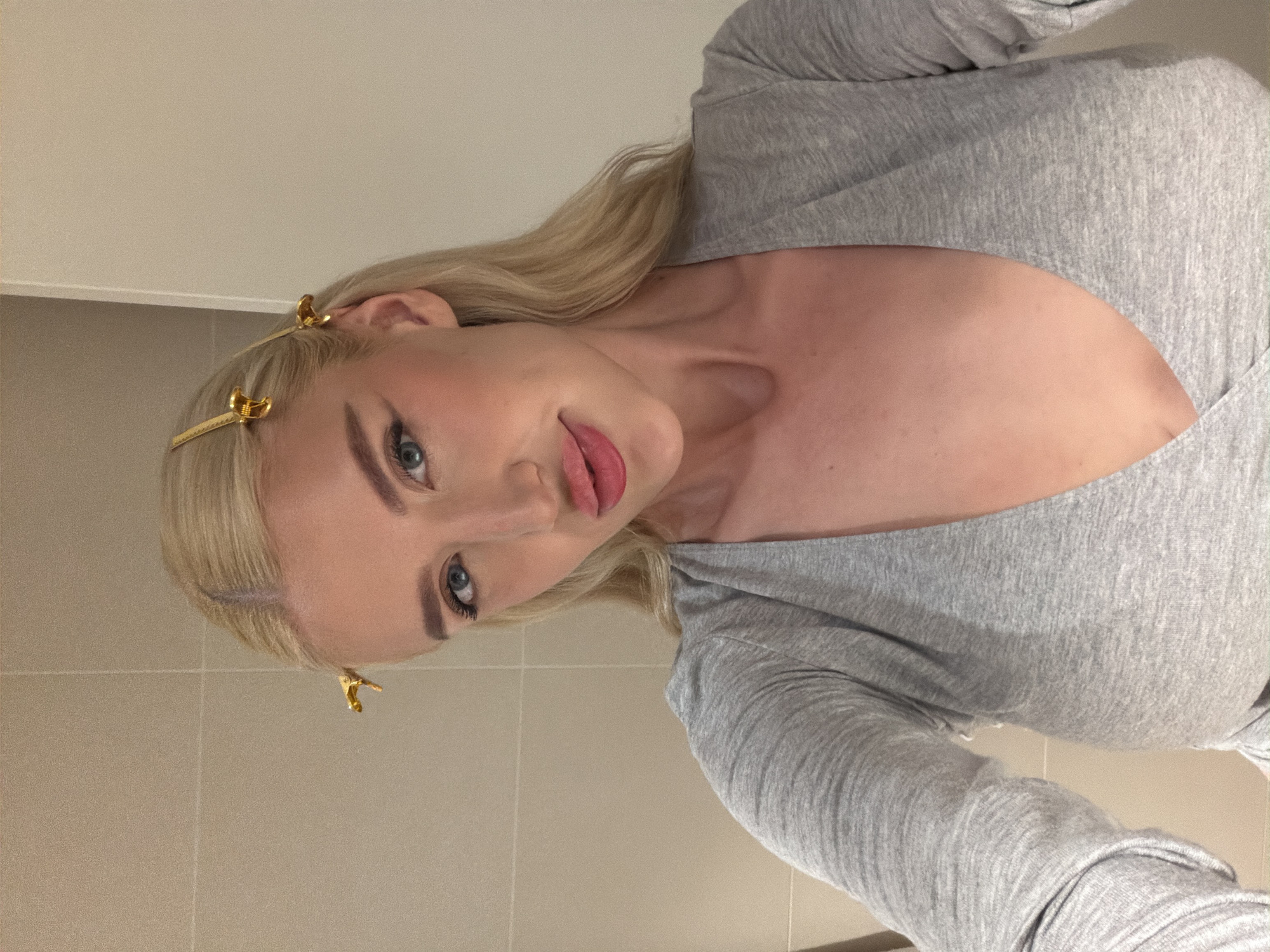 A blonde-haired woman with fair skin and light blue eyes is facing the camera while wearing a gray long-sleeved shirt. She has gold-colored hair clips on the side of her head. The background shows a tiled wall. There is no verifiable information that reveals the city of Belgrade, Serbia in this image.