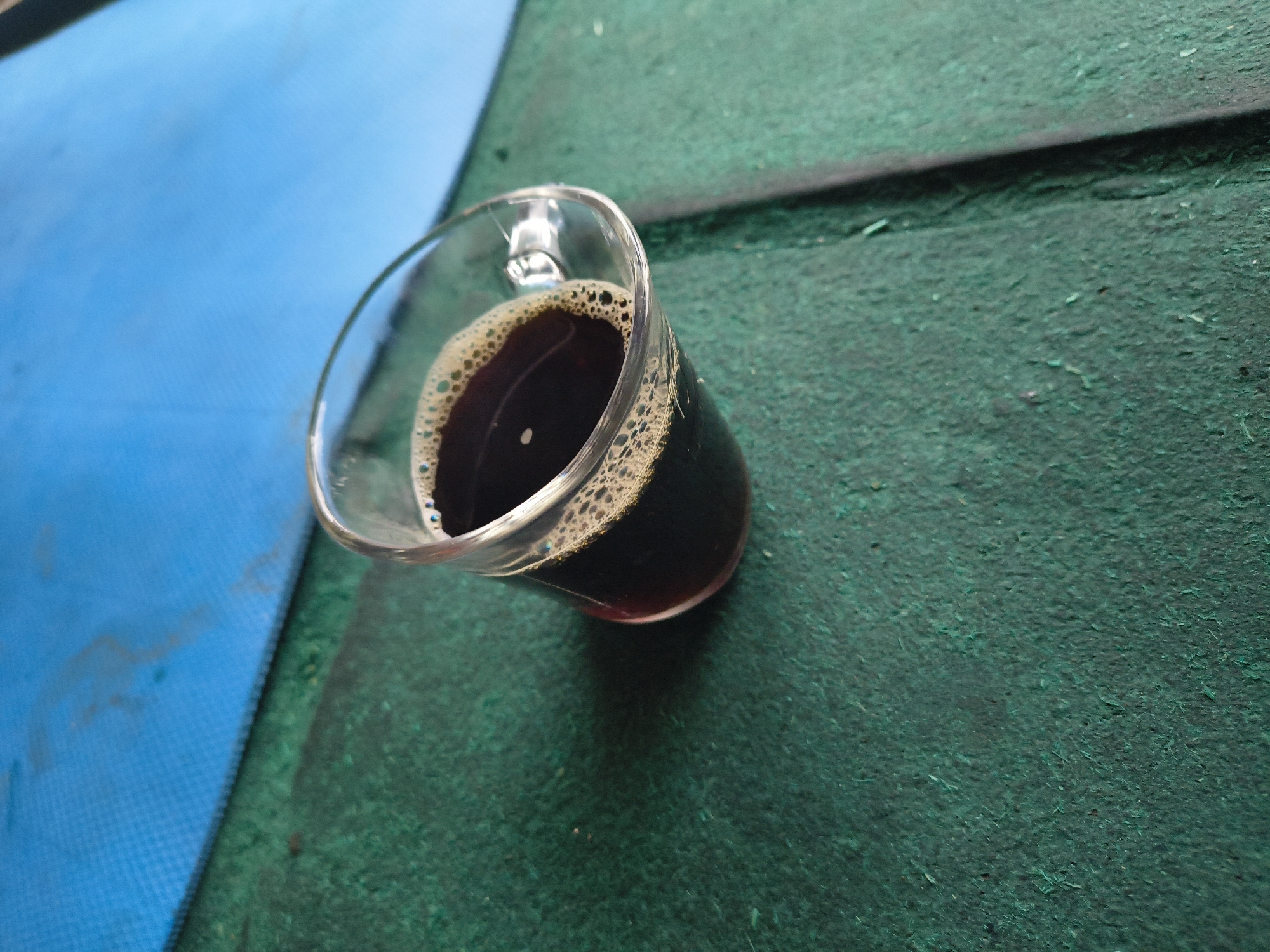 A clear glass cup filled with a dark liquid, likely coffee, sits on a textured green surface adjacent to a blue one. The coffee has a light brown foam layer on top. The image was taken in Bhagsu, India.