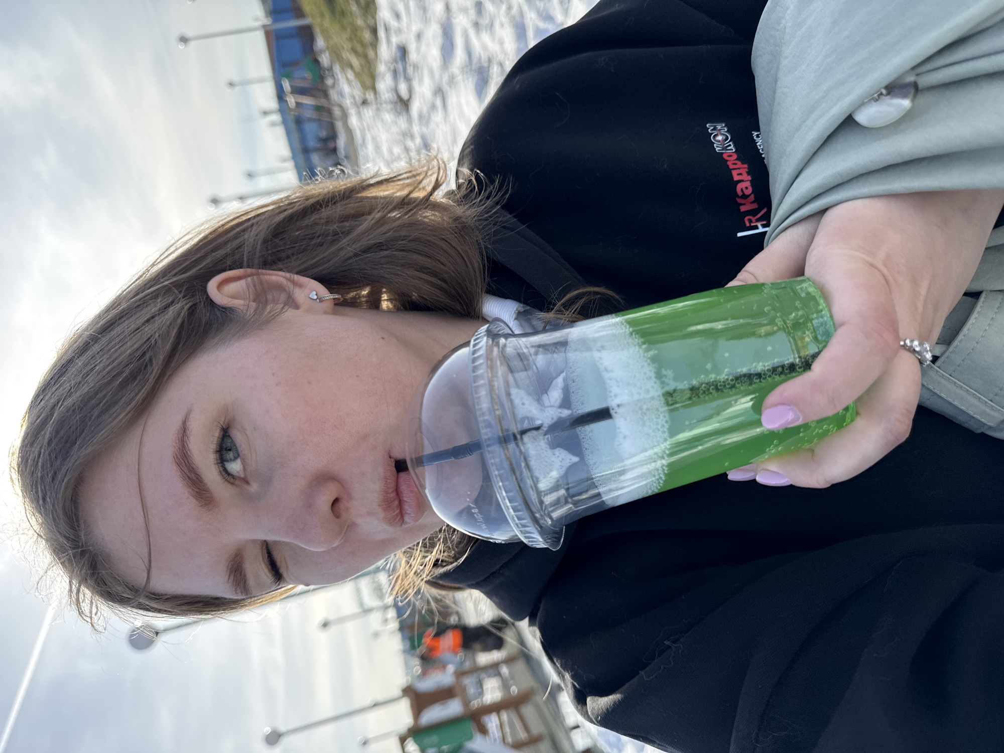 A young woman is holding a clear plastic cup with a green, bubbly beverage and a black straw. She is drinking from the straw. The cup has a clear, domed lid. The woman has light skin, light brown hair, and is wearing a black hoodie. Her nails are painted a light pink color. She is also wearing a light green jacket with a silver button. A ring is visible on her ring finger. The text "FR KAPKOMOH AGENCY" is printed on the sleeve of her hoodie.
The background shows an outdoor setting with snow on the ground and light-colored skies, possibly indicating winter or early spring. There are street lamps and a blue structure in the distance. Based on the writing on the hoodie, it is possible that the location is Perm, Russia, since the text appears to be in Russian Cyrillic.