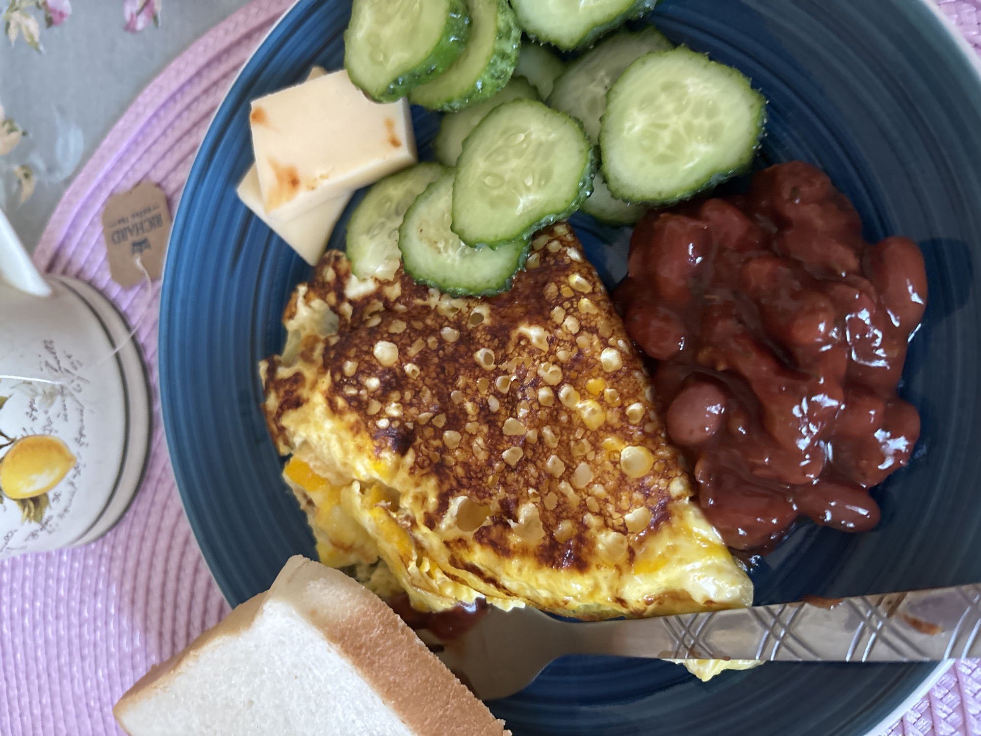 A close-up, top-down view of a blue plate with a breakfast on it, placed on a pink placemat. The breakfast consists of a slice of white bread, a folded omelet with browned edges, a serving of baked beans, sliced cucumbers, and two cubes of cheese. A silver fork is partially visible on the left side of the plate. In the background, a white teacup with a lemon illustration is partially visible. A tea bag tag from "Richard" is also visible. The setting appears to be a domestic environment, possibly a kitchen or dining area. There is no indication of the city of Naberezhnye Chelny, Russia, in the image.