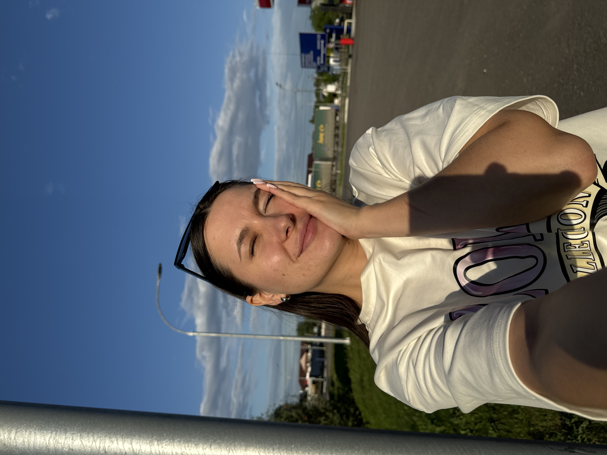 A young woman is holding her hand to her face, eyes closed, against a bright blue sky with scattered clouds. Her sunglasses are perched on her head. She is wearing a white t-shirt with a large graphic design. In the background, there is a parking lot, green grass, and a building. Street signs are visible in the distance. The location appears to be in Bolshiye Vyazyomy, Russia.