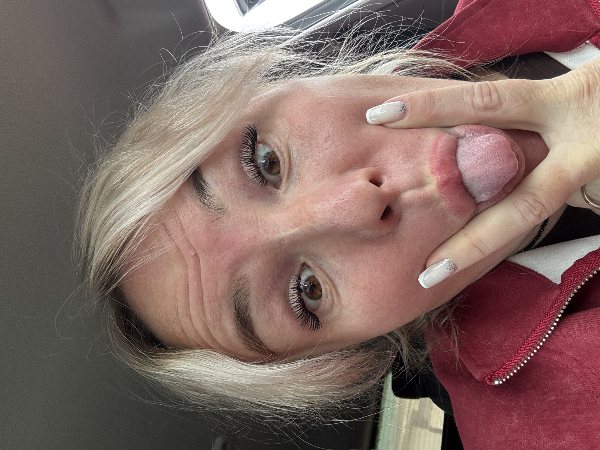 In a close-up, indoor shot, a woman with blonde hair sticks out her tongue and makes a funny face. Her eyes are wide, and her eyelashes are long and dramatic. She's wearing a red jacket, and her fingernails are painted white with glitter. The background is blurry, but it appears to be the interior of a car, suggesting she's on the go. There's no indication of the city of Chastyye, Russia, in this image.
