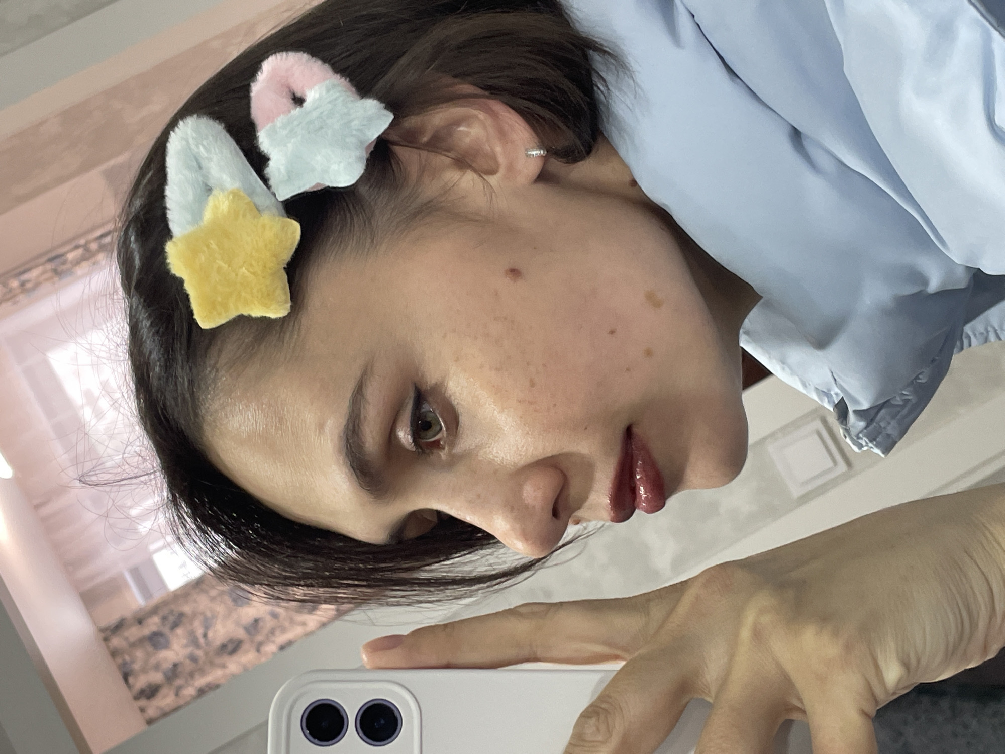A woman with short dark hair is taking a selfie with a white smartphone. She has two colorful hair clips on the side of her head. She is wearing a light blue jacket or shirt. Her left hand is holding the phone. The background shows a partially visible window with a patterned curtain and a white wall. There is no information indicating that the photo was taken in Izhevsk, Russia.