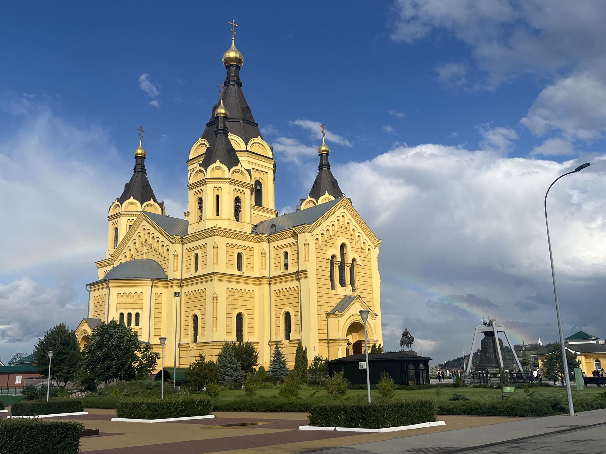 The image captures the exterior of the Alexander Nevsky Cathedral in Nizhny Novgorod, Russia. The predominantly yellow cathedral features multiple black domes topped with golden crosses. In the foreground, manicured green bushes and pathways are visible. To the right, a modern streetlamp stands tall against a backdrop of a dramatic sky filled with both blue patches and dark, heavy clouds. A faint rainbow arcs across the sky, partially obscured by clouds. In the middle ground, a large bell hangs within a metallic frame. A statue of a man on horseback is situated to the left of the bell. Several people are present in the middle ground, walking around the grounds and near the bell structure. The overall scene suggests a recent rain shower followed by clearing skies, indicated by the rainbow and the mix of light and shadow.