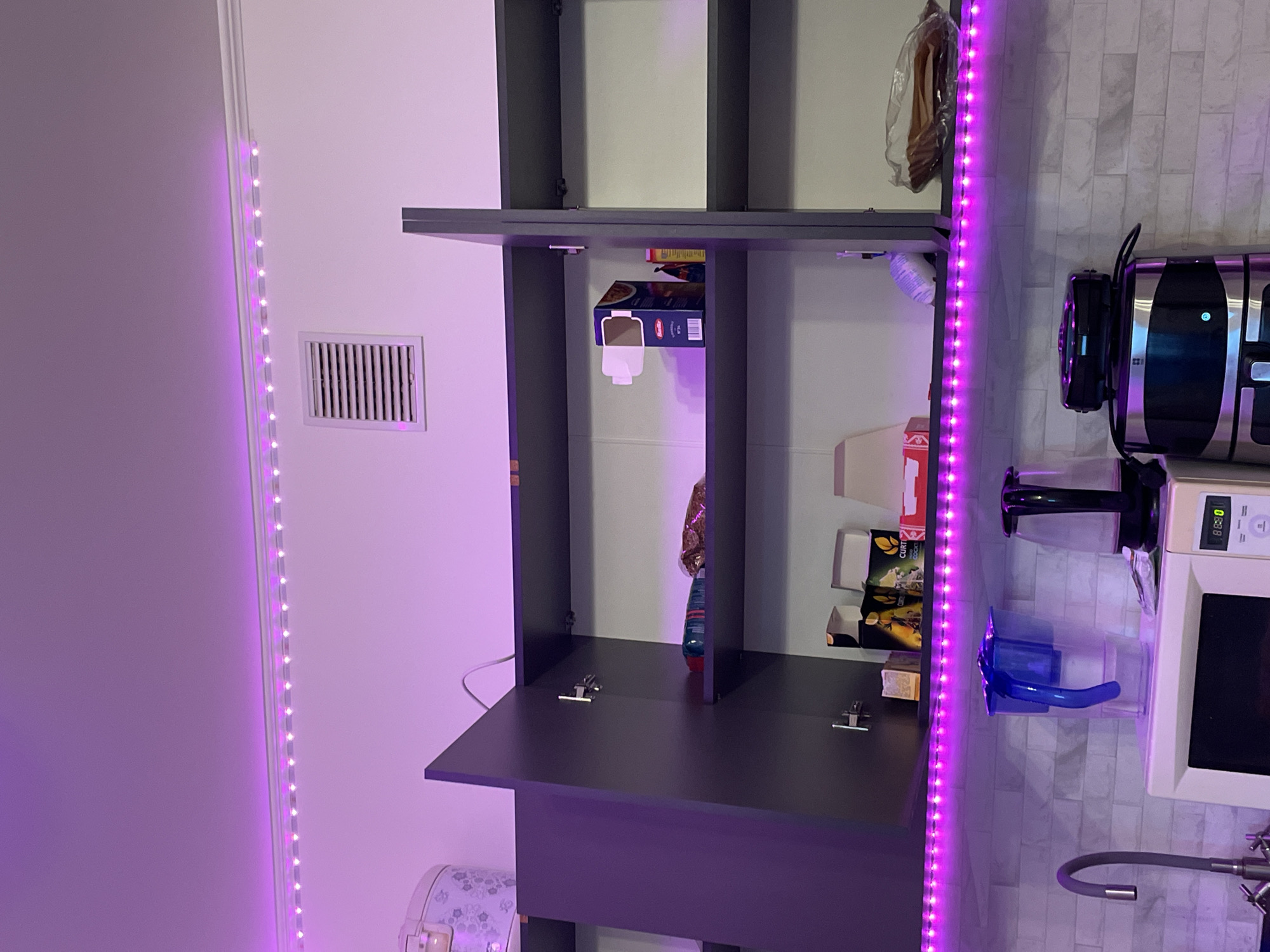 This is a photograph of an apartment kitchen in Ladozhskoe Ozero, Russia. The kitchen features a set of open overhead cabinets, illuminated by purple LED strip lights mounted along the top and bottom edges of the cabinets. The cabinets contain various food items and boxes. Below the cabinets, a white tiled backsplash is visible. On the counter are a microwave, a water filter pitcher, and a glass electric kettle. A kitchen faucet is also visible on the left side of the image. The overall scene suggests a domestic living space.