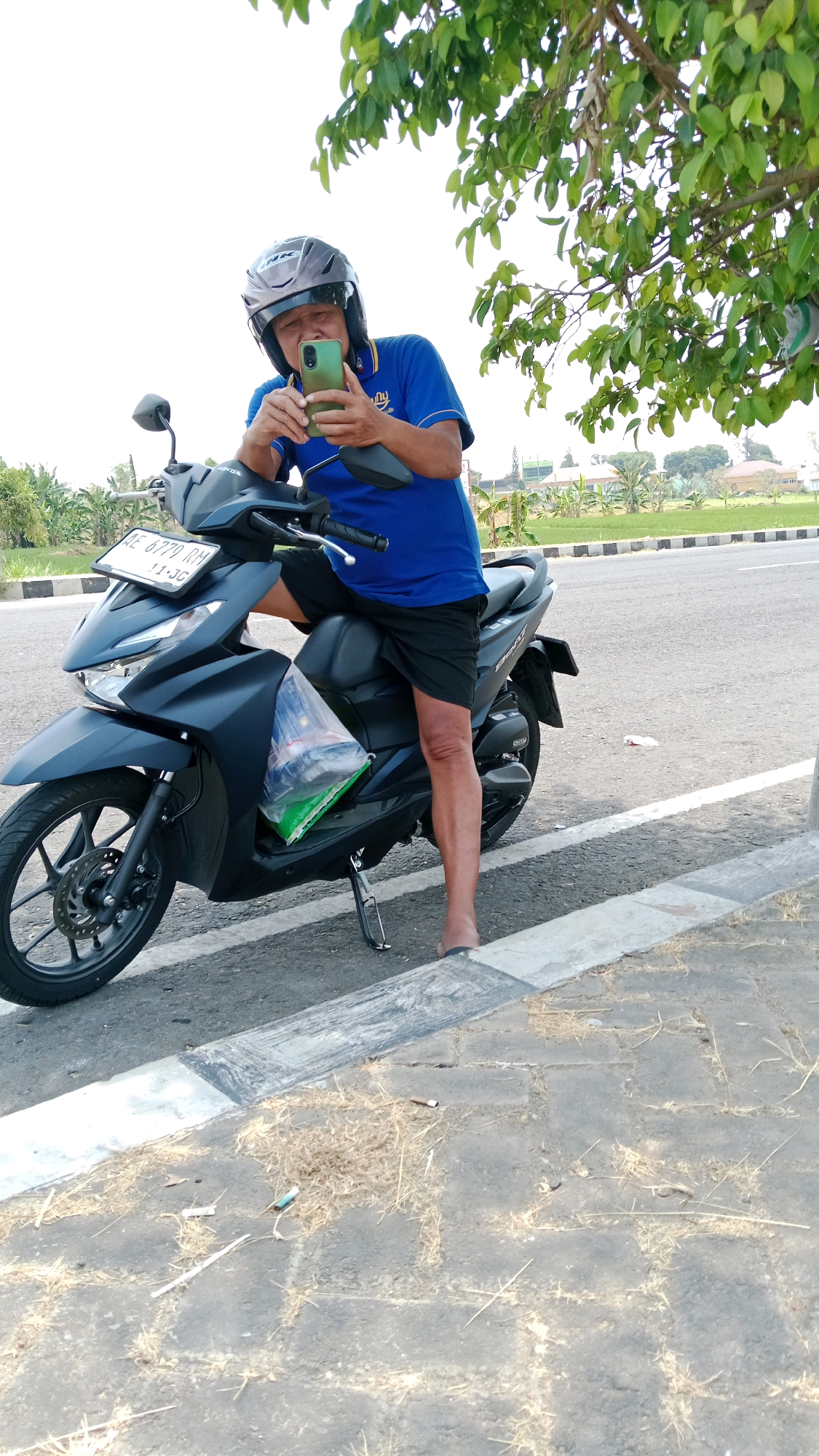 An Indonesian man wearing a blue shirt and black shorts sits on a dark-colored scooter on the side of a road. He is wearing a grey helmet and holding a green smartphone, appearing to take a photo or video. A plastic bag containing items is strapped to the front of the scooter. The scooter has a license plate that reads "BE 6770 RM 11-JG". The man is barefoot, wearing dark flip-flops. The scooter is parked on a paved area next to a road with white markings. In the background, there are fields, trees, and buildings. The sky is overcast and bright. The scene is located in Sukowinangun, Indonesia.