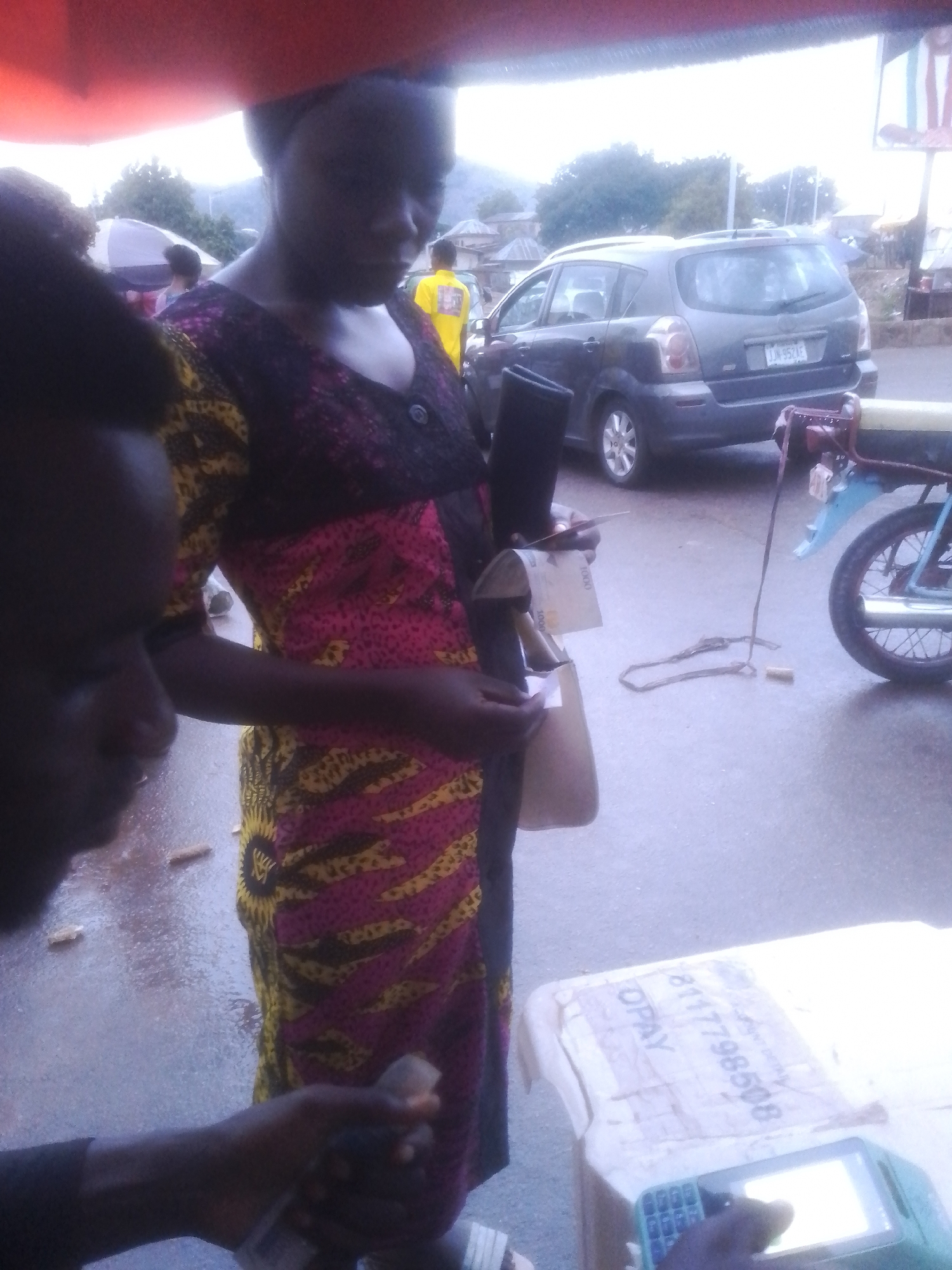 In Gwong, Nigeria, an outdoor scene captures a woman in a vibrant pink and yellow patterned dress, holding a small tan bag and what appears to be money. Her expression is neutral as she engages with a mobile payment device, a teal-colored terminal with a lit screen and a keypad, resting on a white surface. Another person, partially visible on the left, with dark hair and skin, seems to be interacting with the payment process, holding a phone or similar device.
In the background, a dark grey hatchback car, possibly a Toyota, is parked, its license plate reading "LJN 95Z AE." A blue and yellow motorcycle is also present, partially obscuring the view of the car. A man in a bright yellow shirt stands further back, near the car. The setting suggests a street or roadside location, with a glimpse of buildings and trees in the distance. The ground is wet, indicating recent rain. The time of day appears to be late afternoon or early evening, with soft, diffused light and a somewhat overcast sky. The overall atmosphere is one of everyday commerce and activity. Visible text on the payment terminal includes "OPAY" and a string of numbers "8117798508." Some of the currency held by the woman also shows the numeral "1000."