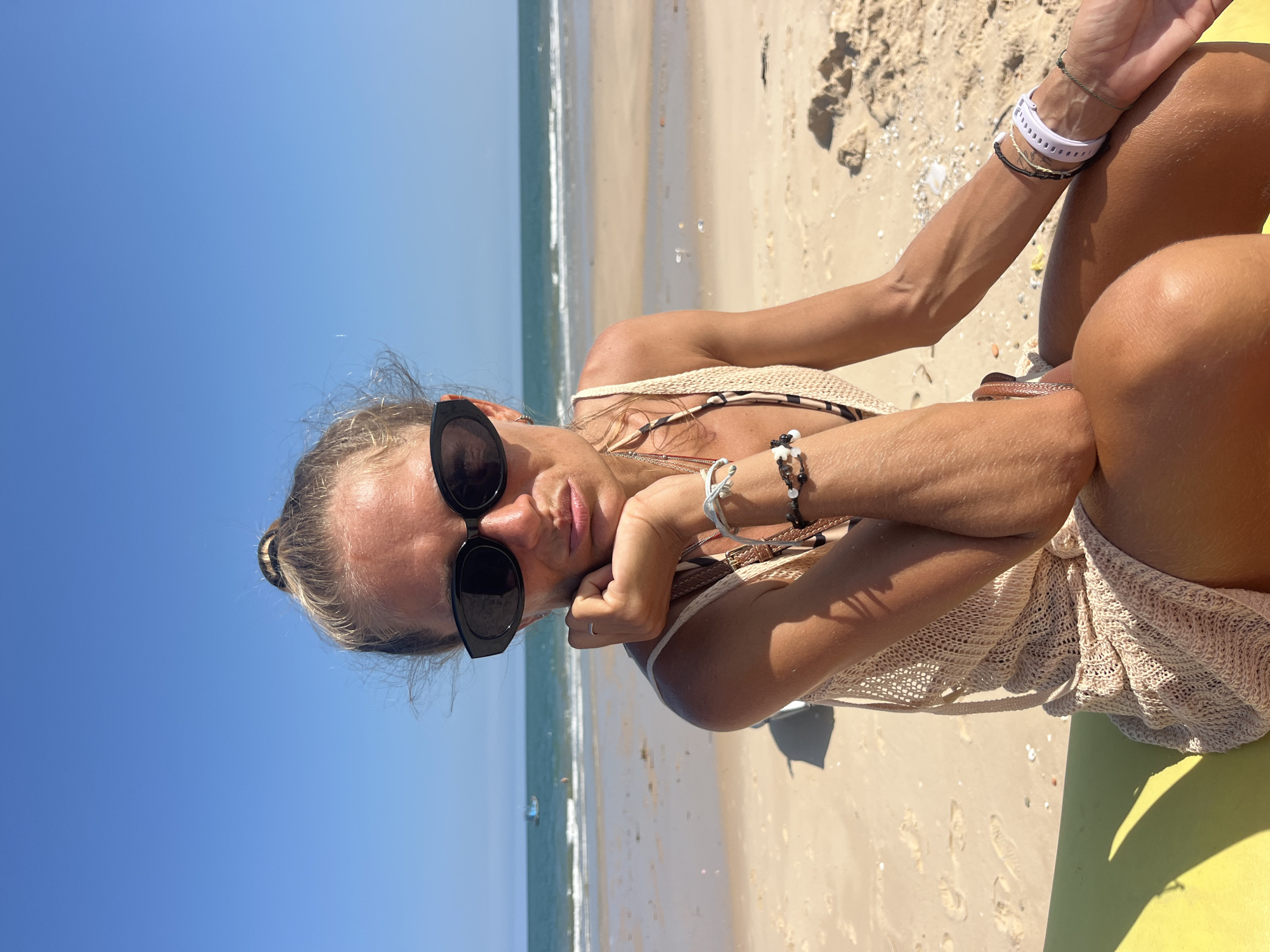 A woman with blonde hair pulled into a bun, wearing large black sunglasses and a beige knitted cover-up over a patterned bikini, sits on a yellow surface on a sandy beach. She rests her chin on her hand, looking at the camera with a pout. Behind her, the ocean meets a clear blue sky. Footprints are visible in the sand, and a small boat is in the distance on the water. The scene appears to be in Phu Thuy Ward, Vietnam.