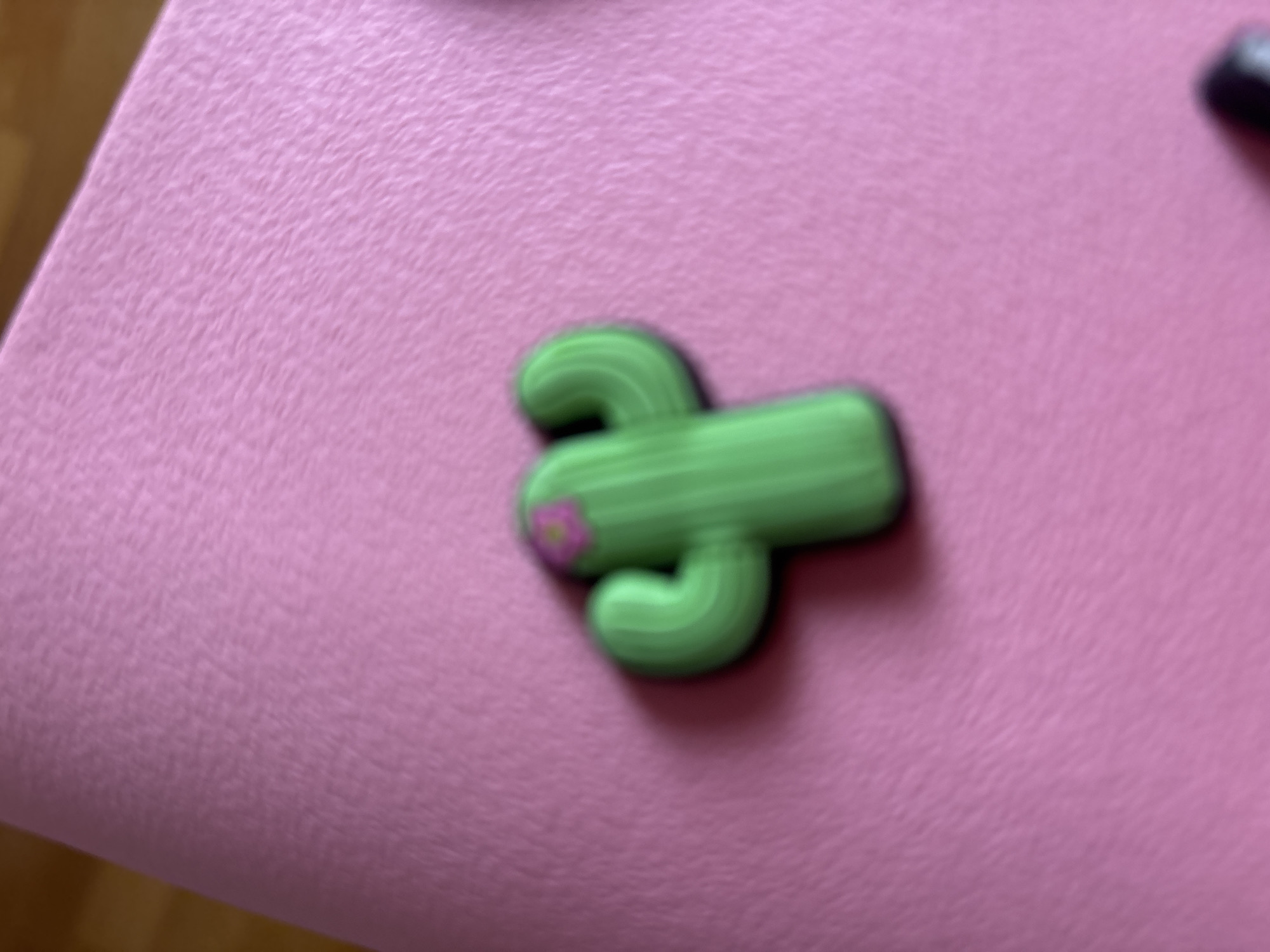 A green cactus-shaped charm with a pink flower detail is positioned on a textured pink surface. The charm is blurred, indicating motion or a shallow depth of field. There are no people or other identifiable objects in the immediate vicinity of the charm. The background is out of focus, with a hint of a wooden surface in the upper left corner.