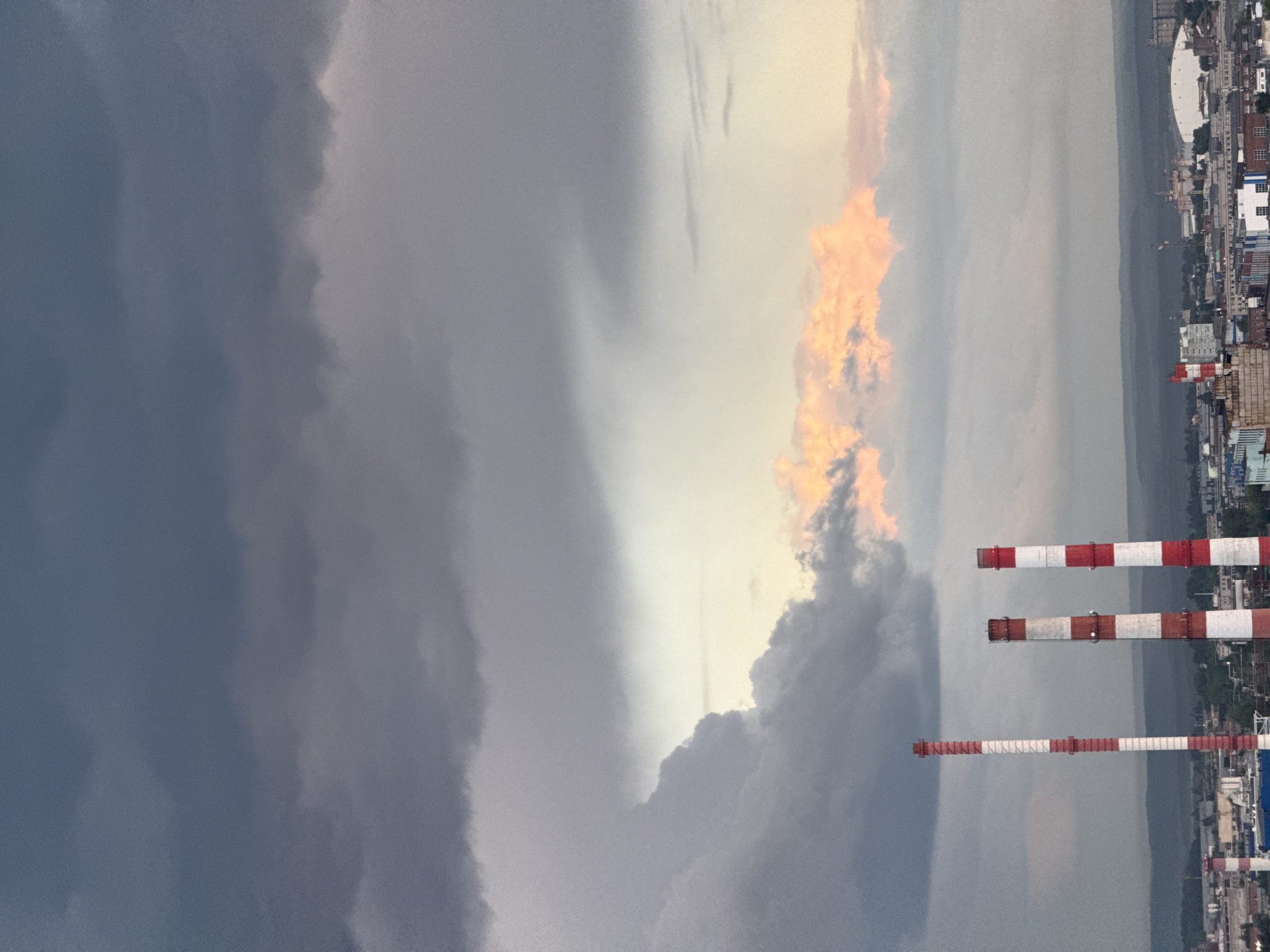 This is a medium-distance, landscape, outdoor shot taken from an elevated viewpoint looking over a city skyline under a dramatic sky. In the foreground, three tall, industrial smokestacks with red and white alternating bands dominate the frame. The leftmost smokestack is partially visible, while the other two are fully in view. The smokestacks are positioned in front of a sprawling city with a mixture of low-rise and mid-rise buildings, including some industrial structures and a large domed building on the right side of the frame. In the far background, rolling hills or mountains are visible. The sky is a prominent feature, filled with dark, heavy storm clouds that transition to lighter grays and whites towards the horizon, with a patch of warm, peachy-colored clouds illuminated by the sun peeking through in the middle distance. The overall mood is one of impending weather change. There are no people visible in the image.