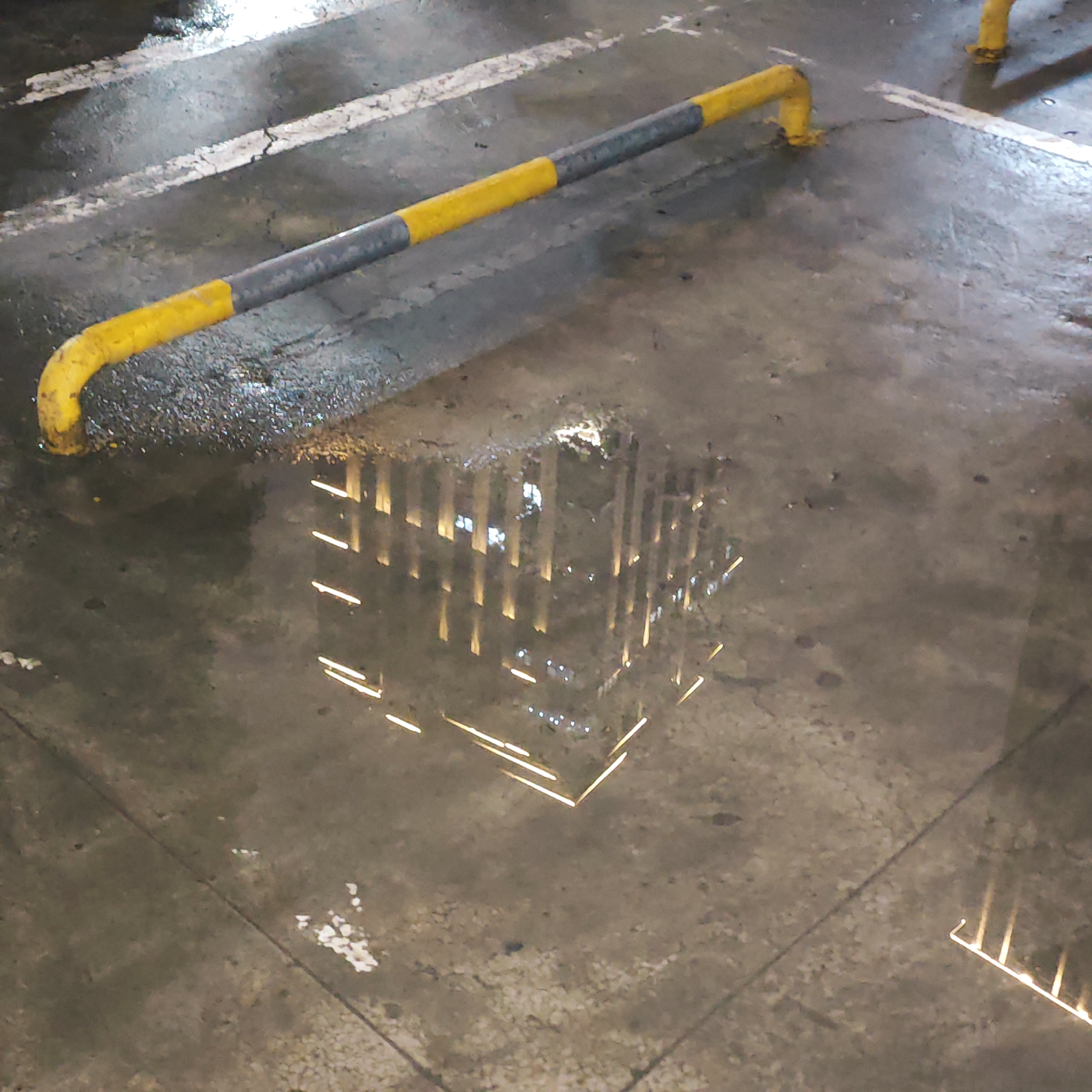 This nighttime image, set in Yuzhong District, China, captures a wet concrete ground, likely an outdoor urban space such as a parking lot or alleyway. The primary subject is a large puddle reflecting a towering building. The reflection shows numerous illuminated windows and sections, creating a luminous, almost ethereal appearance for the high-rise structure. The building appears to be tall, possibly a residential or commercial skyscraper.
Diagonally across the upper half of the frame, a yellow and dark gray striped metal barrier, resembling a protective railing or parking stop, is prominent. Its reflective surface indicates the presence of ambient light. The ground itself is textured with visible cracks, wear, and patches of white painted lines, suggesting an aged and utilitarian surface. The overall wetness implies recent rain, contributing to the reflective qualities of the scene. No people or specific activities are visible, and the scene is devoid of any discernible text. The image conveys a quiet, almost contemplative mood, emphasizing the interplay of light and reflection in an urban environment after rainfall.