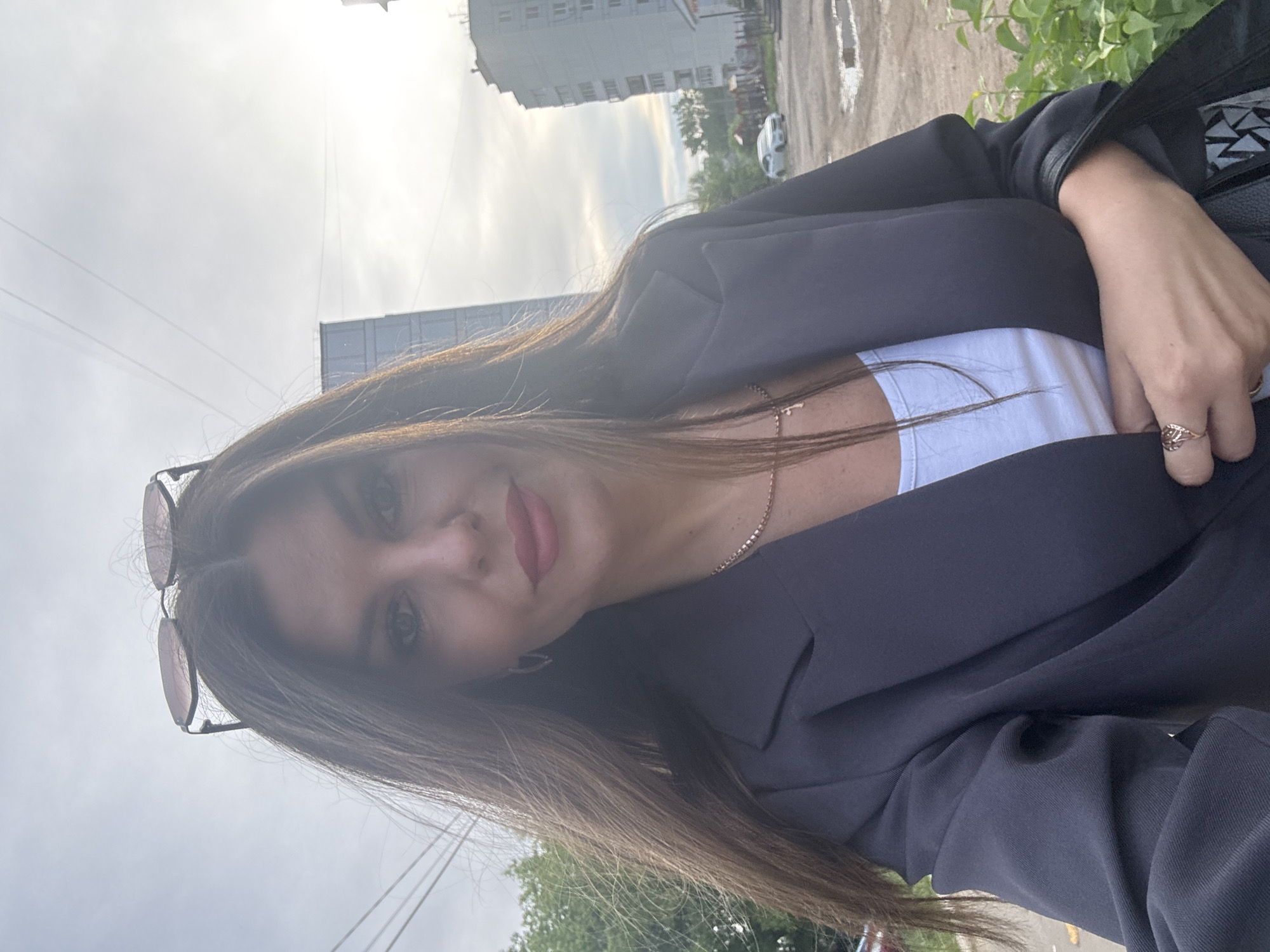A young woman is pictured outdoors, likely in Chaikovsky, Russia. She's wearing a dark blazer over a white top, and eyeglasses are propped on her head. She's also wearing jewelry, including a necklace and ring. The background reveals a city scene with buildings, foliage, and a roadway with puddles, suggesting recent rain. The cloudy sky indicates overcast weather conditions.