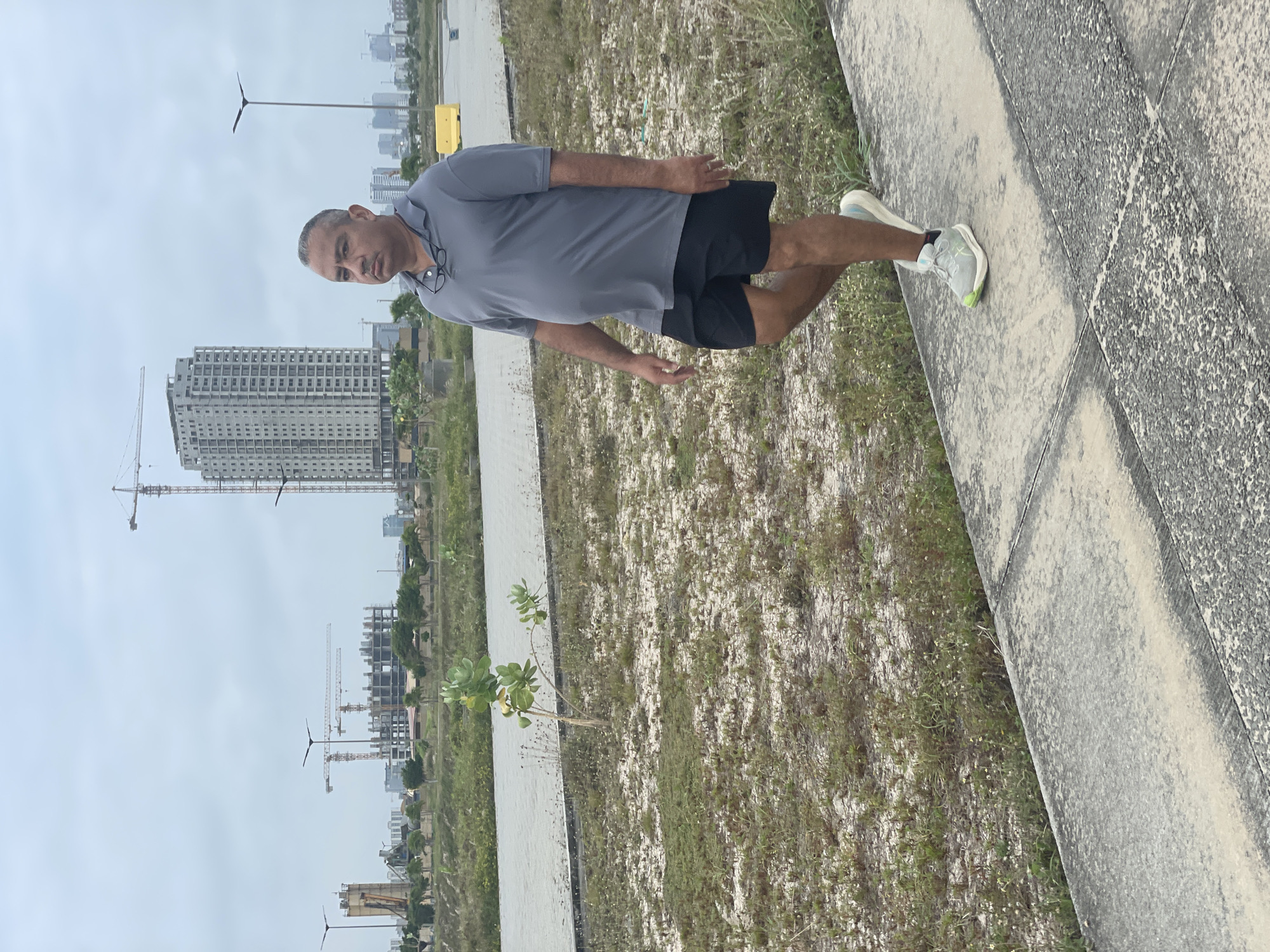 A man walks on a paved path in what appears to be a developing urban area, possibly Inupa Kekere, Nigeria. The man, who is of average build and appears to be middle-aged, is wearing a grey polo shirt, black shorts, and running shoes. He is looking towards the right of the frame with a neutral expression. Behind him, a tall, unfinished building dominates the skyline, with several construction cranes visible in the background, suggesting ongoing development. The surrounding landscape is sparsely vegetated with dry grass and small trees, and a body of water is visible in the mid-ground. The sky is overcast, casting a diffused light over the scene.