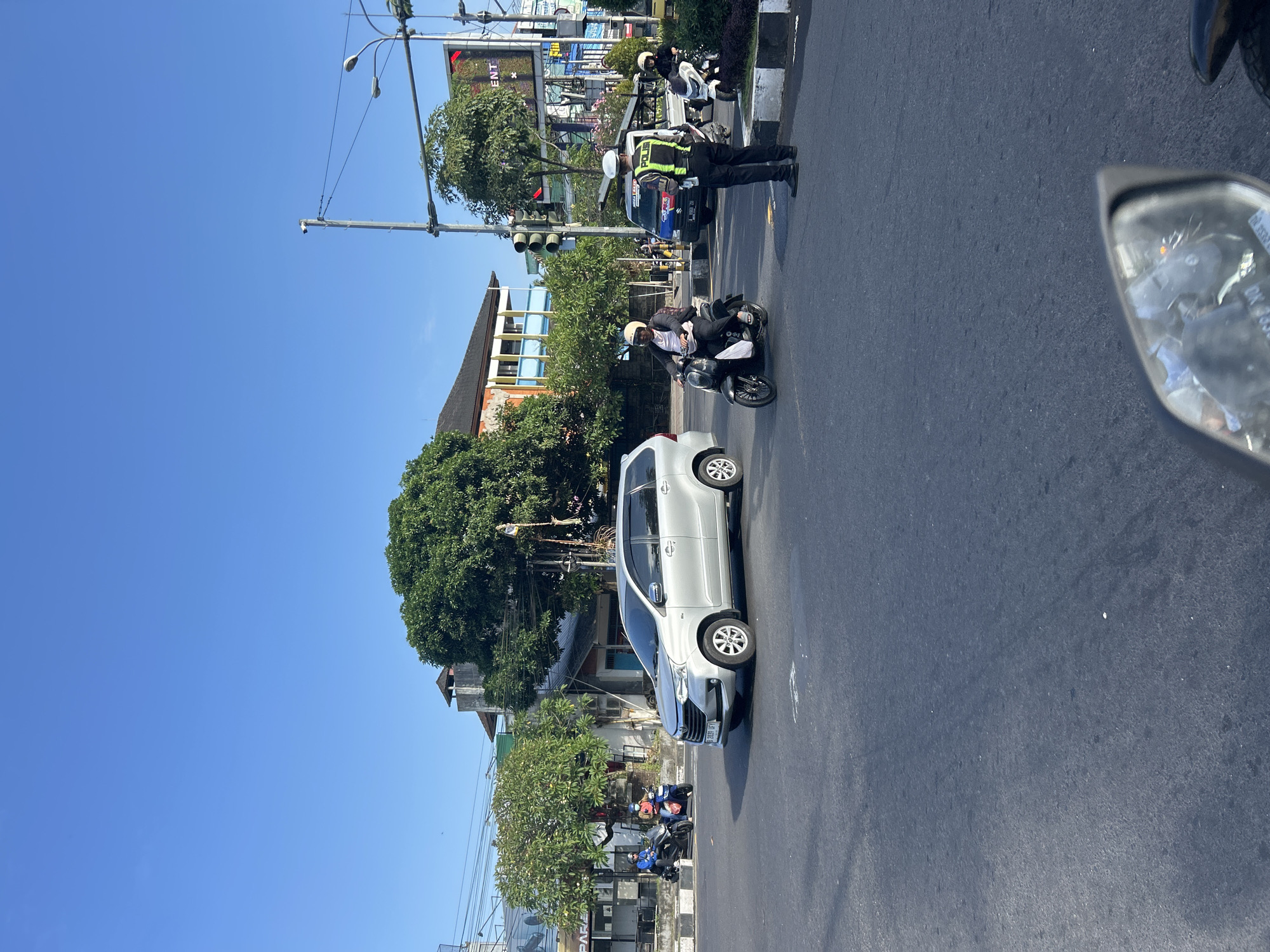 The image depicts a street scene in Denpasar, Indonesia, likely an intersection on a clear day. A silver minivan is stopped at the intersection, with a person on a dark scooter approaching from the right. A police officer in uniform, wearing a reflective vest and a white helmet, stands in the middle of the road, directing traffic. Several other scooters and a light truck are visible in the background, along with buildings and trees. The overall environment suggests a busy urban area.
