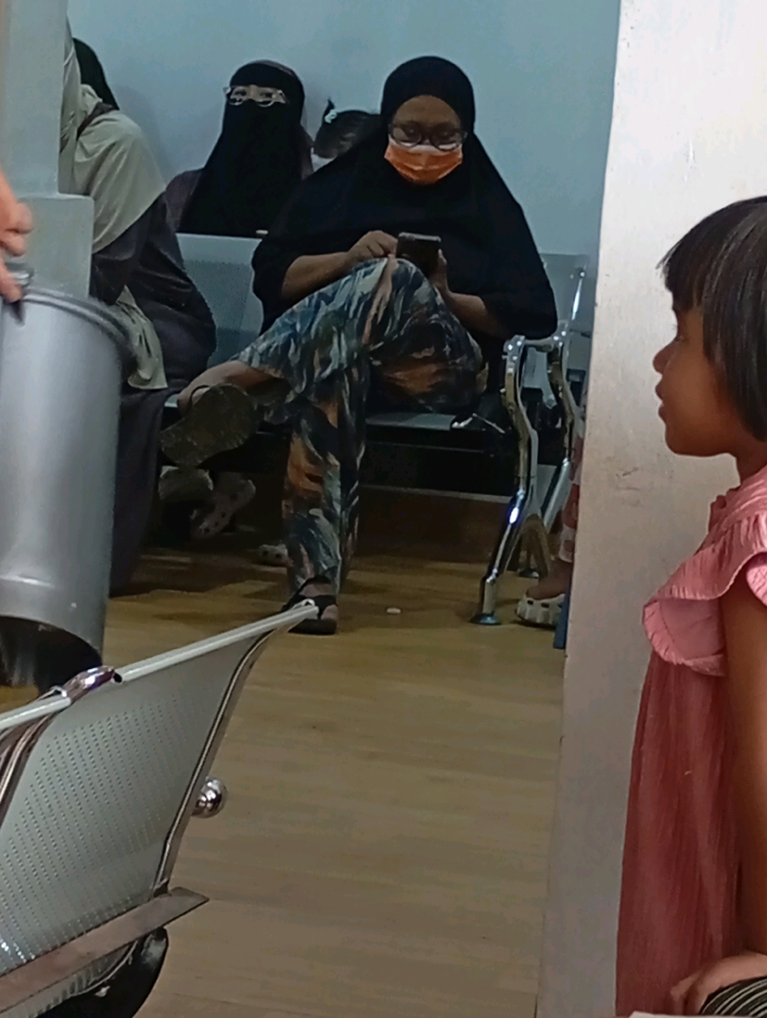 In a waiting room, likely a public health facility in Babelan, Indonesia, a woman wearing a black niqab and orange face mask, and glasses, is seated and looking at her phone. Her patterned pants are visible as she leans forward. To her left, another woman in a light-colored hijab and dark clothing is also seated. In the foreground, a young girl with dark hair, wearing a pink ruffled top, is partially visible on the right, facing away from the camera. The room has light-colored walls and a wooden floor. In the mid-ground, chrome chairs and a large silver cylindrical object are present. The scene suggests a typical waiting area in a busy location.