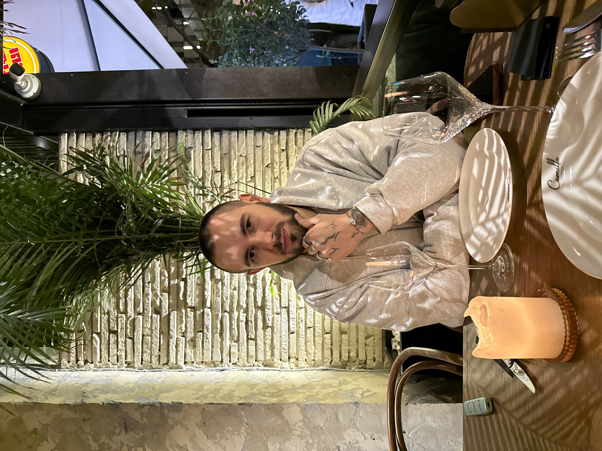 A man with short hair and tattoos sits at a restaurant table, wearing a gray hooded sweatshirt. He is looking at the camera. On the table are two plates, wine glasses, silverware, and a candle. The restaurant has a brick wall and is decorated with plants. Based on the plate, the location is Yerevan, Armenia.