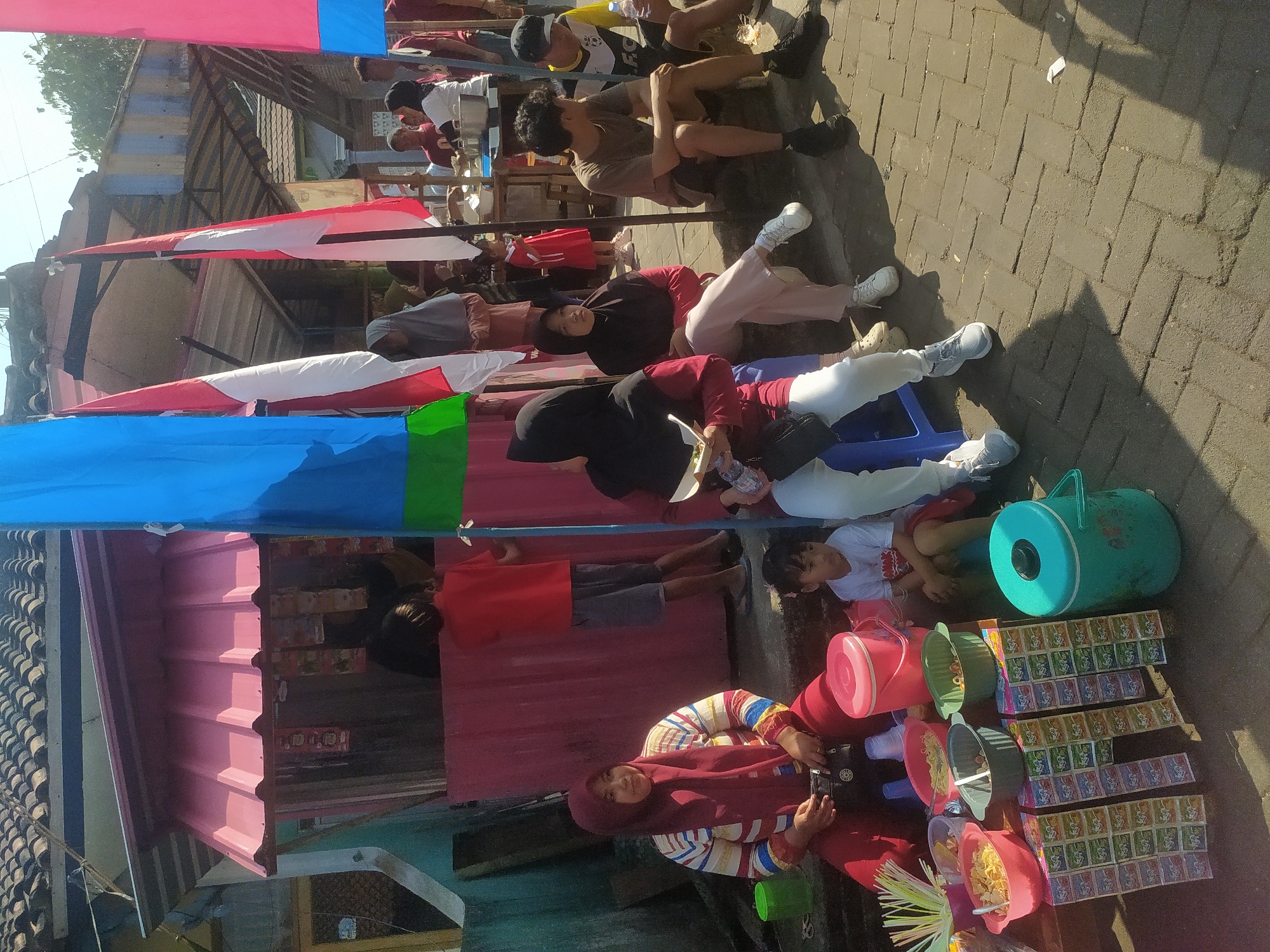 A group of people are gathered around a food stall on a paved street in Lopait, Indonesia. The stall has a pink roof and displays various food items and snacks. Several individuals are seated or standing, some appear to be customers and others vendors. Flags are displayed prominently, adding to the festive atmosphere. The weather appears to be sunny, casting shadows on the ground.