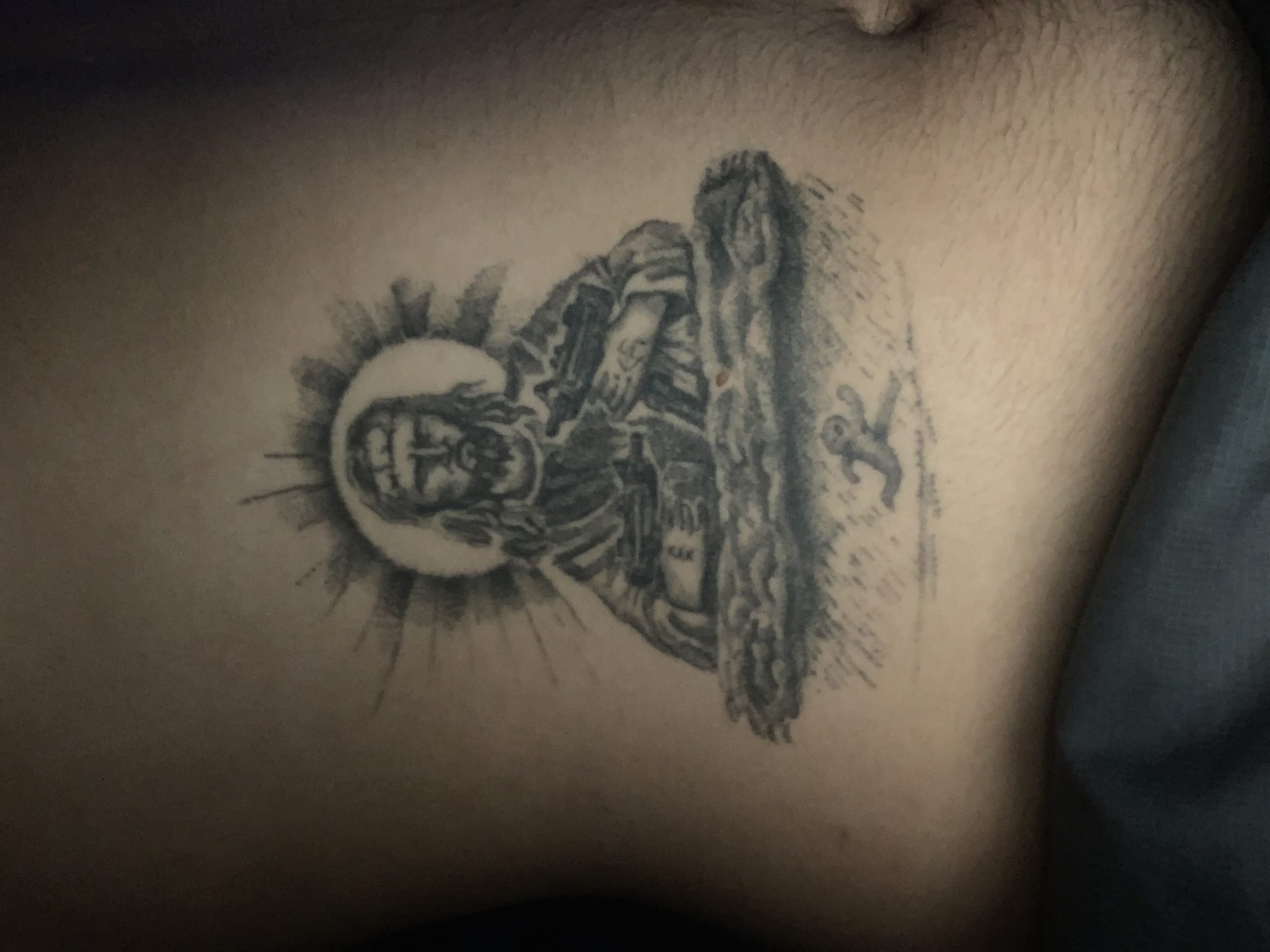 The image shows a tattoo on human skin. The tattoo depicts a religious figure, possibly Jesus Christ, with a halo. The figure is positioned next to a stylized tree or wooden structure, with a small, abstract image that could be a bird or symbol next to it. The skin is fair and has some hair. The location in Buenos Aires, Argentina is unknown.