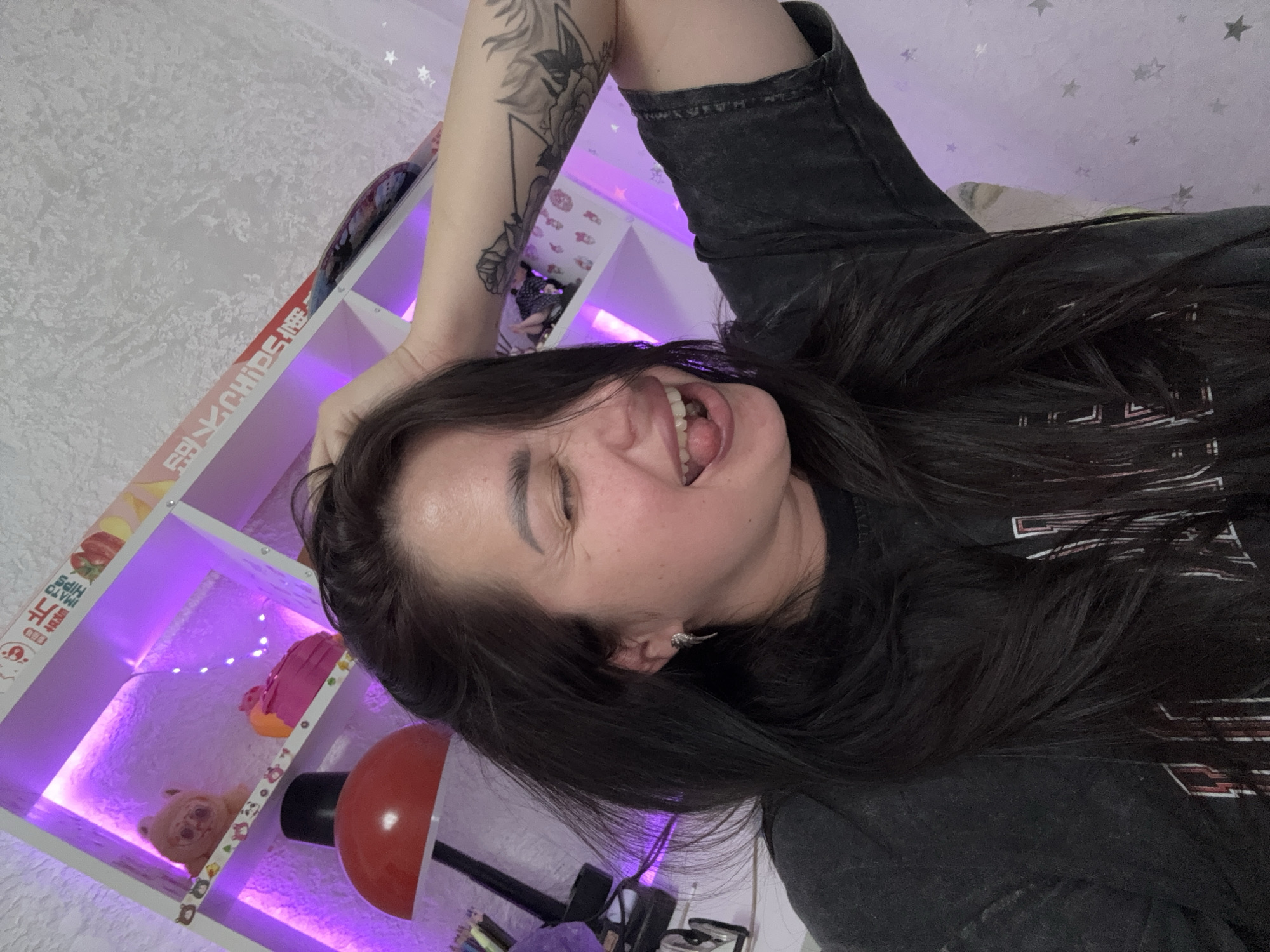 A woman is shown from the chest up, posing in front of a shelf with purple LED lighting. She has dark hair and is wearing a dark t-shirt. Her right hand is raised, touching her hair, and her eyes are closed with her mouth open, sticking out her tongue. She is wearing a silver earring. The shelf behind her contains various items, including stuffed animals and decorative objects. The background wall has a textured pattern with small star shapes. There is a red lamp visible to the left of the woman. The scene appears to be indoors, possibly a bedroom or personal space. There is no indication of the image being taken in Tolyatti, Russia.