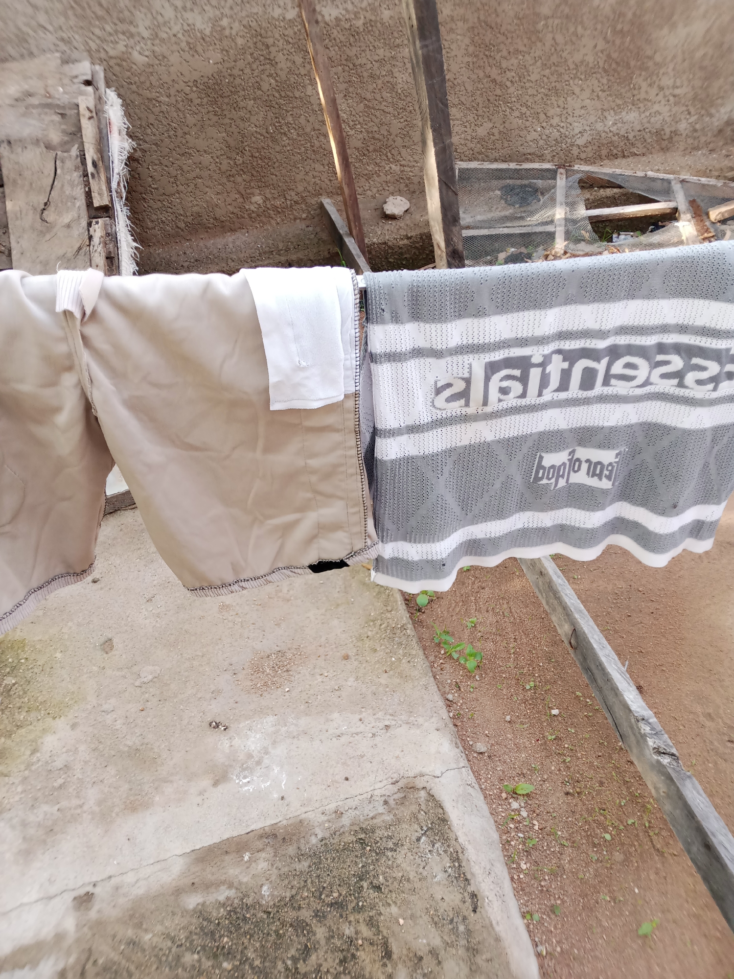 The image shows clothing hanging on a clothesline in what appears to be an outdoor setting in Jauro Yinu, Nigeria. The subjects are two pieces of clothing: on the left, a pair of tan shorts or pants, and on the right, a gray and white patterned fabric, possibly a blanket or a towel, with visible text. The shorts are made of a smooth fabric with stitching along the hem and a waistband. The patterned fabric has horizontal stripes and a diamond-like pattern in gray and white. Two lines of text are visible on this fabric. The top line appears to be "efitnes922" or a similar arrangement of letters and numbers, possibly a brand name or slogan. Below this, a smaller line of text reads "boplotag".
The scene is outdoors with a concrete ground and a dirt area with sparse vegetation. In the background, there are wooden structures and a rough, textured wall or ceiling, suggesting an area that is partly covered or under construction. The lighting suggests it is daytime. The overall impression is one of a casual, domestic scene, likely a laundry area. There are no people visible.