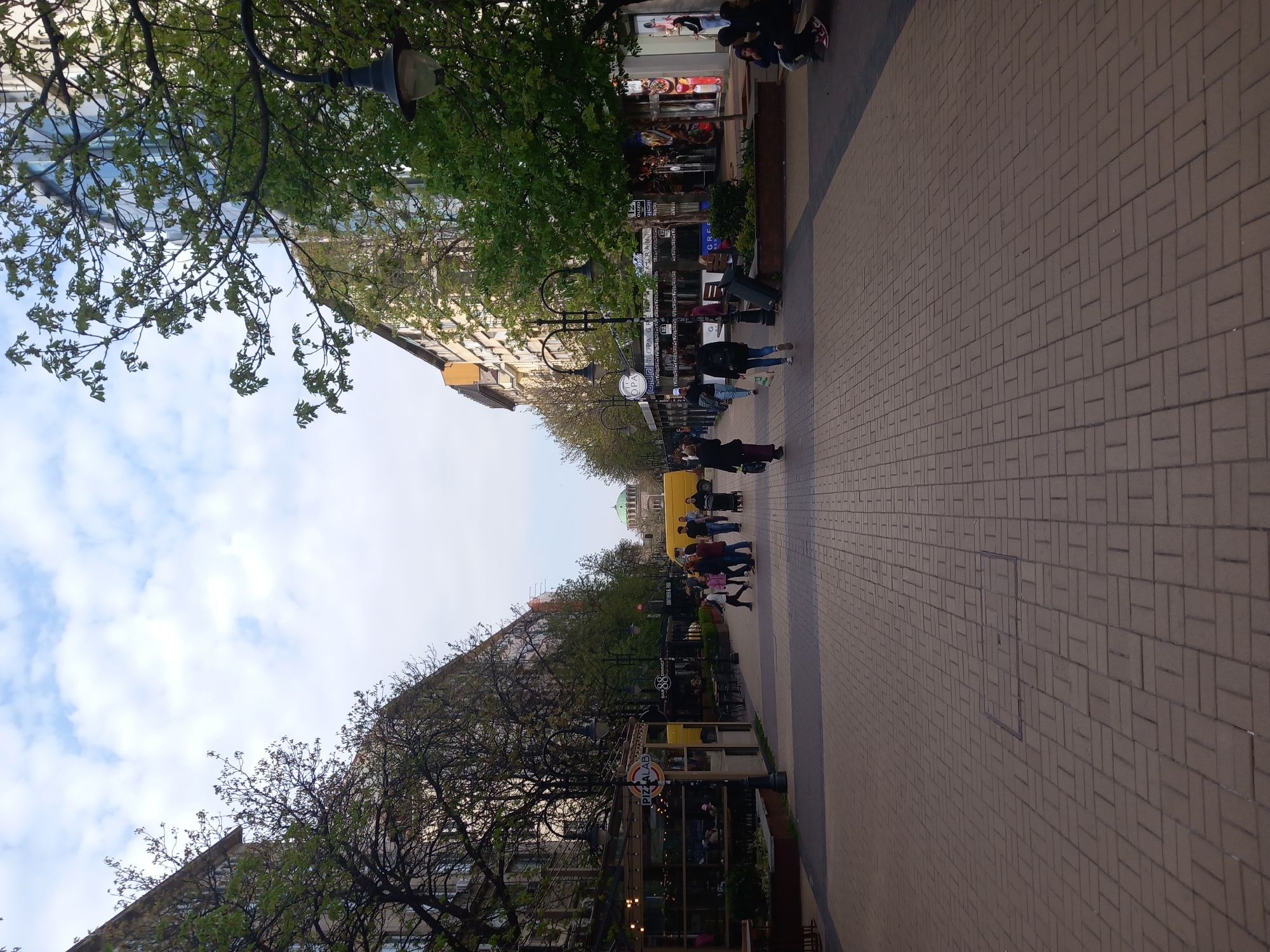 The image shows a pedestrian street in Sofia, Bulgaria. The street is paved with rectangular bricks and lined with buildings on both sides. Trees are planted along the street, providing shade. People are walking along the street in both directions. A clock tower is visible in the middle of the street. A yellow truck is parked on the street. There are outdoor dining areas for restaurants along the street. The sky is partly cloudy. In the distance is visible the dome of a church.