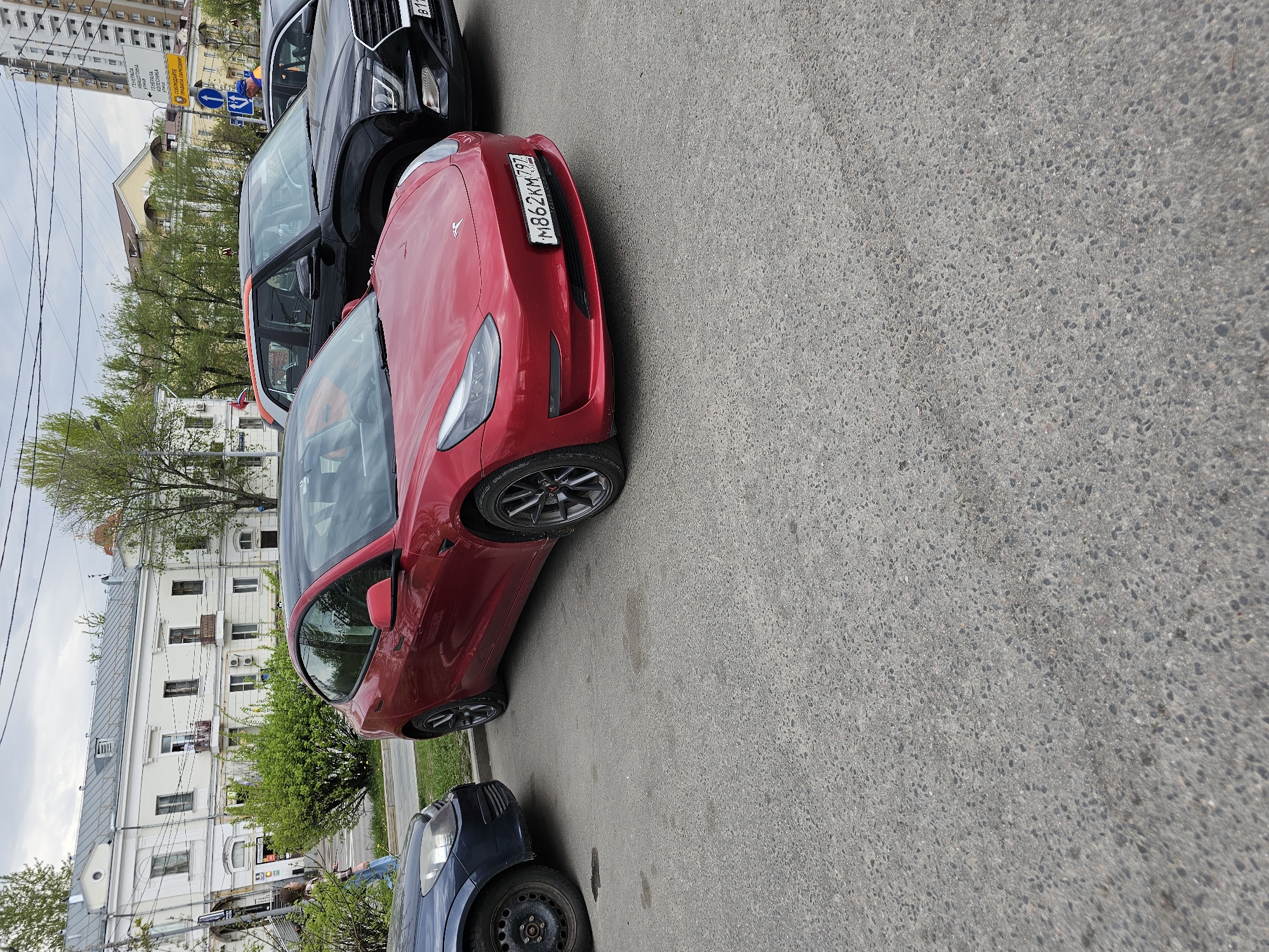 The image depicts a street scene in Moscow, Russia, featuring a red Tesla Model 3 parked on the side of the road. The car's license plate reads "M862KM797," indicating registration in the Moscow region. The presence of Russian signage, architecture, and license plates confirms the location. Other vehicles are parked nearby, including a dark-colored SUV. Buildings are visible in the background, along with trees and other street elements like signs and a small green patch. The weather appears overcast.