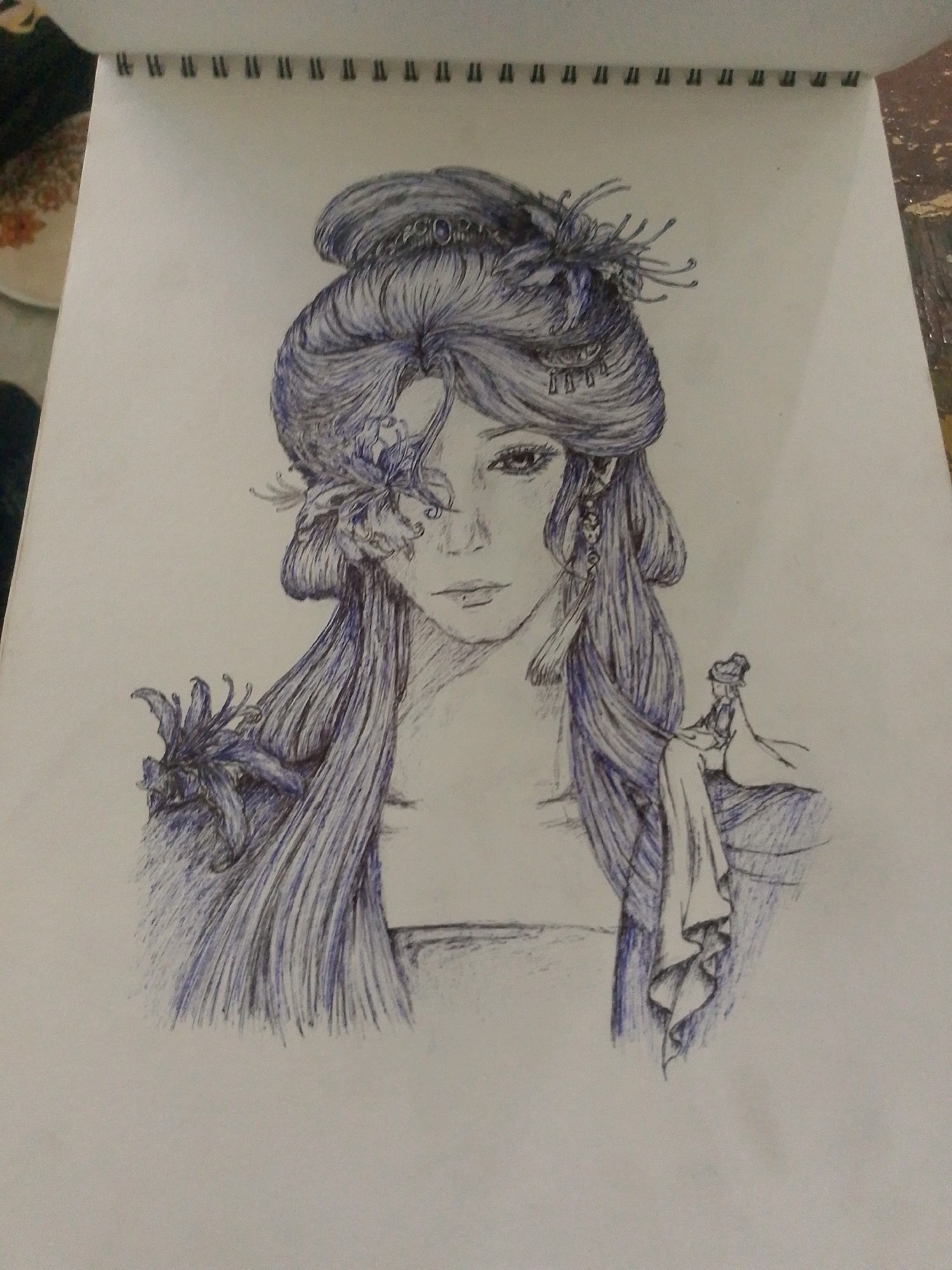A sketch on white paper depicts a woman with intricate braided hair and floral adornments, drawn in blue ink. Her eyes are large and expressive, and she wears elaborate jewelry, including earrings. Her long, flowing hair cascades down her shoulders, adorned with additional flowers. To her right, a smaller figure, possibly a child, is seated, draped in flowing fabric. The sketch is part of a notebook, indicated by the spiral binding at the top. The background is a plain white surface, suggesting an indoor setting. The city of Sitarampuram, India, is not depicted in this image.