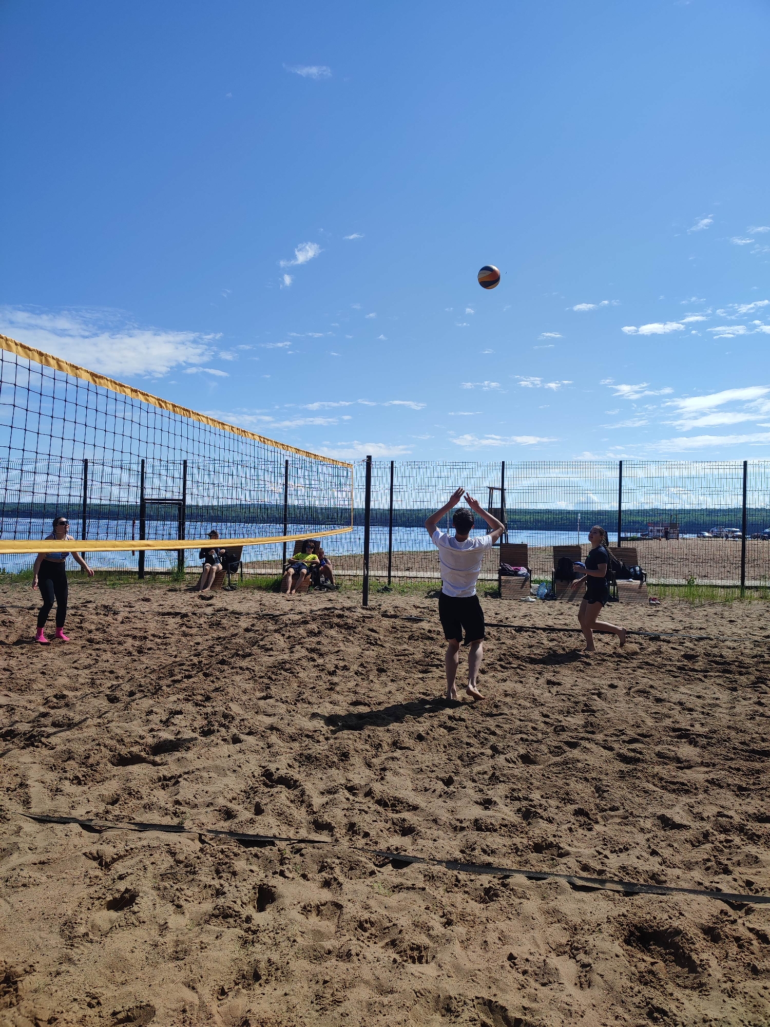 This image depicts an outdoor beach volleyball game under a clear blue sky with scattered cumulus clouds, taking place in the city of Unknown, Unknown. The scene is brightly lit by direct sunlight, casting visible shadows on the sandy court.
The foreground is dominated by a rough, tan sandy surface of a volleyball court, with a dark boundary line visible in the sand. A yellow and black volleyball net stretches across the middle ground, separating the court.
Three primary players are in action on the court. On the left, a woman wearing dark leggings, a light-colored top, sunglasses, and pink shoes stands near the net, looking towards the center of the court. In the center-right, a barefoot man in a white t-shirt and dark shorts is seen from behind, with both arms raised in a setting posture. A multicolored volleyball (orange, white, and blue/black) is suspended in the upper-right quadrant of the sky, indicating active play. To the right of the man, a barefoot woman in a black t-shirt and black shorts is moving across the sand.
Beyond the volleyball court and net, a calm, expansive body of blue water, likely a lake or large river, stretches into the background. A dark metal mesh fence runs along the water's edge, separating the court area from the water and the distant treeline visible across the water. Several wooden beach chairs are positioned on the sand behind the net and near the fence. At least three individuals are seated in these chairs, observing the game, with other personal belongings like bags and water bottles visible nearby.