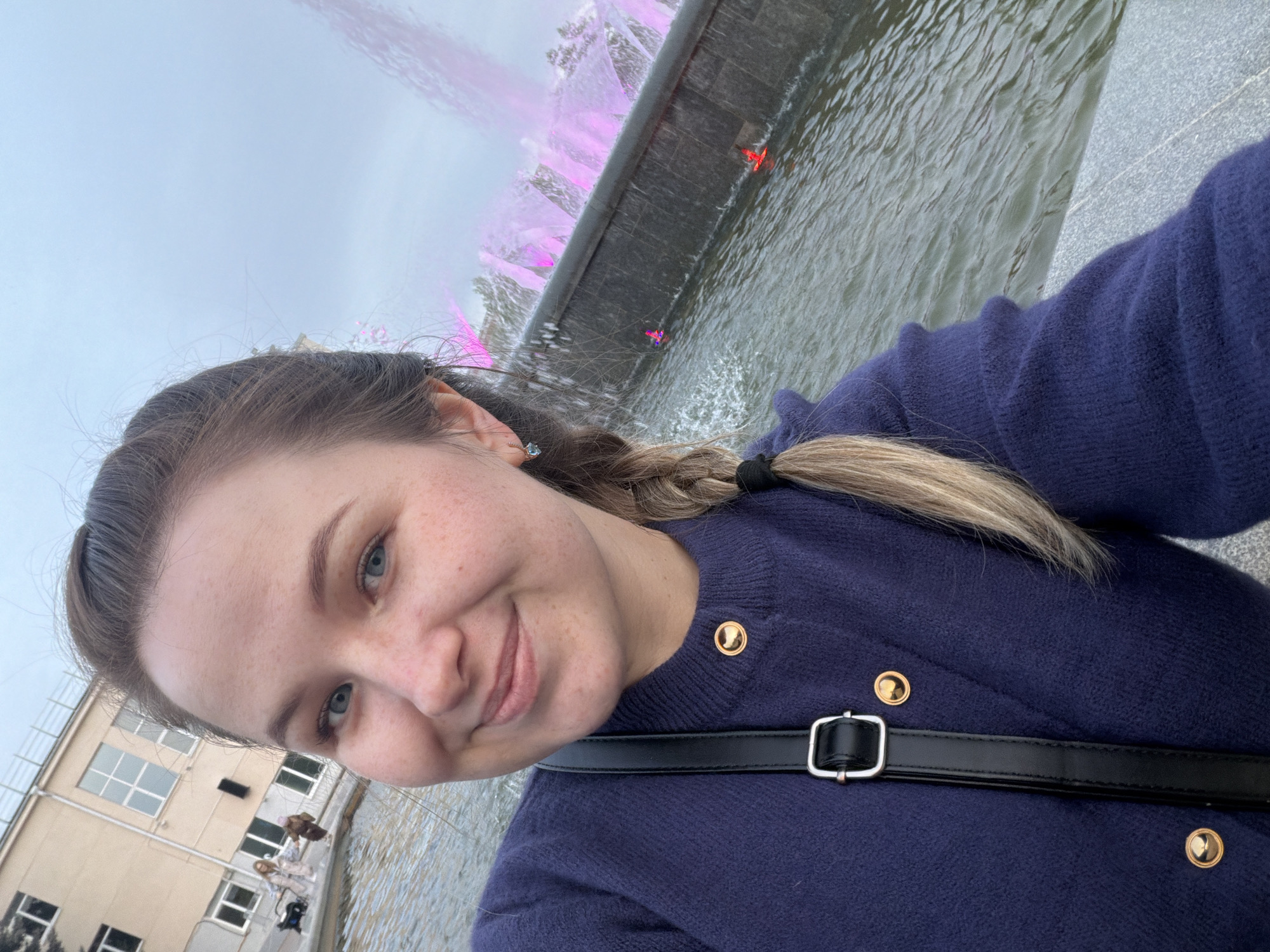 The image is a selfie of a young woman in front of a fountain. She has fair skin, blue eyes, and her hair is pulled back in a braid. She is wearing a dark blue cardigan with gold buttons and a black strap is visible across her chest.
Behind her is a modern-style fountain with water cascading down a dark grey structure. The water is illuminated with pink lighting. In the background, there is a building with multiple windows and a grey sky overhead.
Based on the fountain and building style, this image likely was taken in Chaikovsky, Russia.