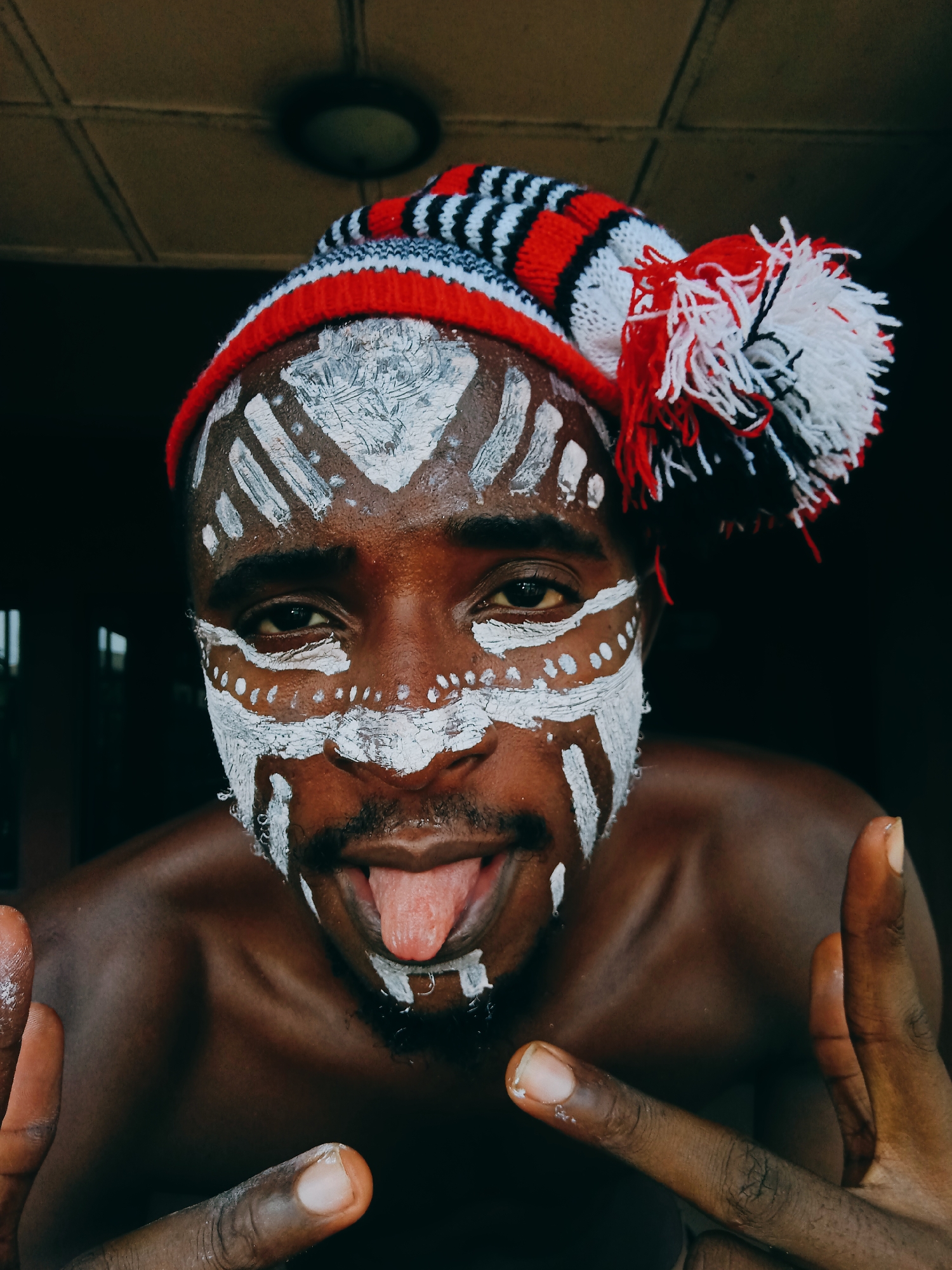 A close-up shot depicts a dark-skinned man with tribal-style white paint on his face, wearing a red, white, and black knitted cap. The man sticks out his tongue and holds up his hands in a gesture of peace. The location is most likely indoors in Nkpor, Nigeria.