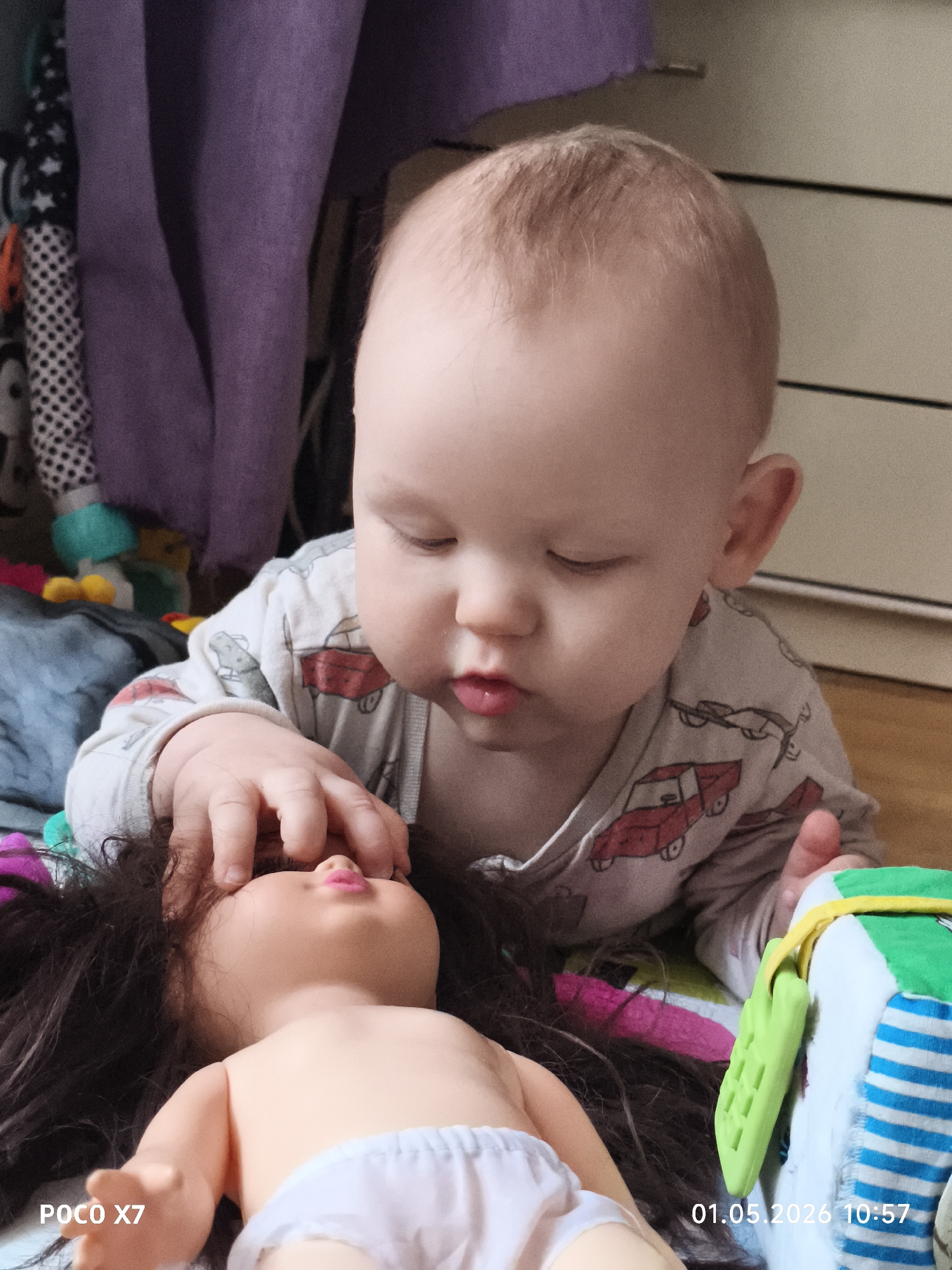 This eye-level indoor photograph, captured in Chaikovsky, Russia, depicts a baby, likely a toddler, engrossed in play. The primary subject is the baby, with light-colored hair and fair skin, lying on their stomach and looking intently at a doll.
The baby is dressed in a light-colored long-sleeved onesie featuring a pattern of red and grey trucks. With a focused and curious expression, the baby gently touches the face of a doll with their right hand, specifically near its eyes. The doll, which has long dark hair and pink lips, is lying on its back, partially on a striped, colorful fabric, and is wearing white underwear.
To the right of the doll, a soft, multi-colored block toy is visible, featuring green, white, and blue stripes, with a bright green textured handle attached. In the background, domestic elements include a folded grey blanket or cloth near the baby, a mass of purple fabric (possibly a curtain or clothing) on the left, and a light-colored wooden or laminate floor extending to a white dresser or cabinet on the right.
The scene conveys a calm moment of childhood exploration during what appears to be daytime. The bottom left corner of the image displays "POCO X7", indicating the camera model, and the bottom right corner shows "01.05.2026 10:57", denoting the date and time the picture was taken.