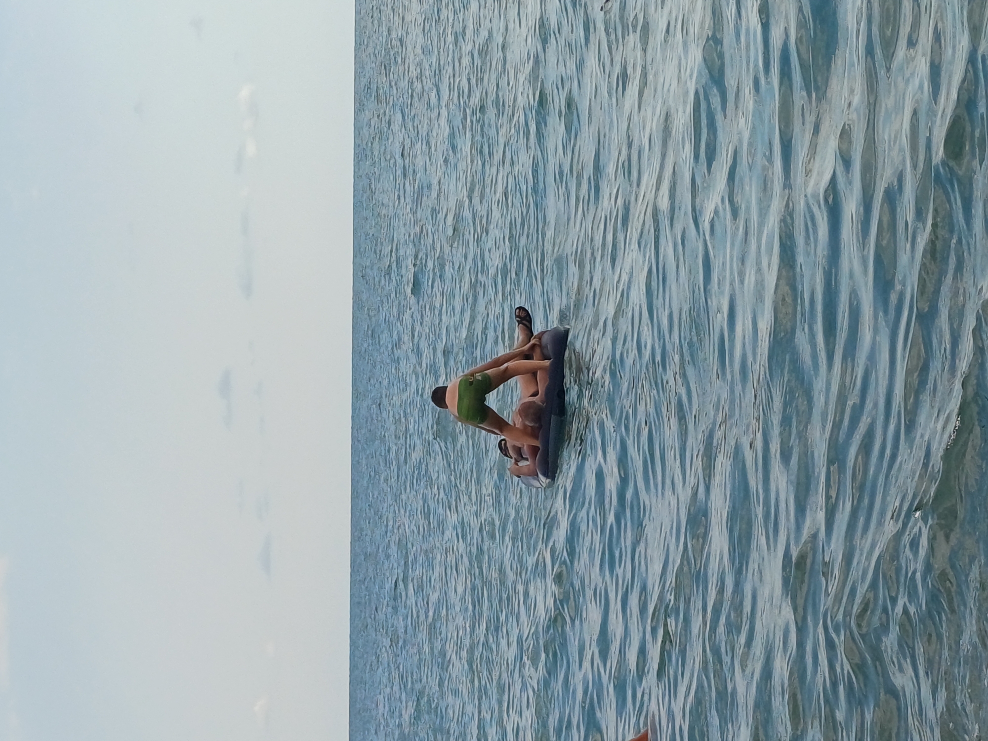 The image depicts two individuals on an inflatable mat in the water. The subject in the foreground is wearing green swim trunks, while the other subject is partially obscured. They appear to be relaxing or enjoying recreational activity on the water. The scene is set on a body of water, likely a lake or the sea, with visible ripples and waves, suggesting movement and possibly a breezy day. The lighting indicates it is daytime, and the sky appears to be overcast or hazy on the left side of the frame. No text is visible in the image. The overall mood seems relaxed and summery, typical of leisure activities at the water's edge.