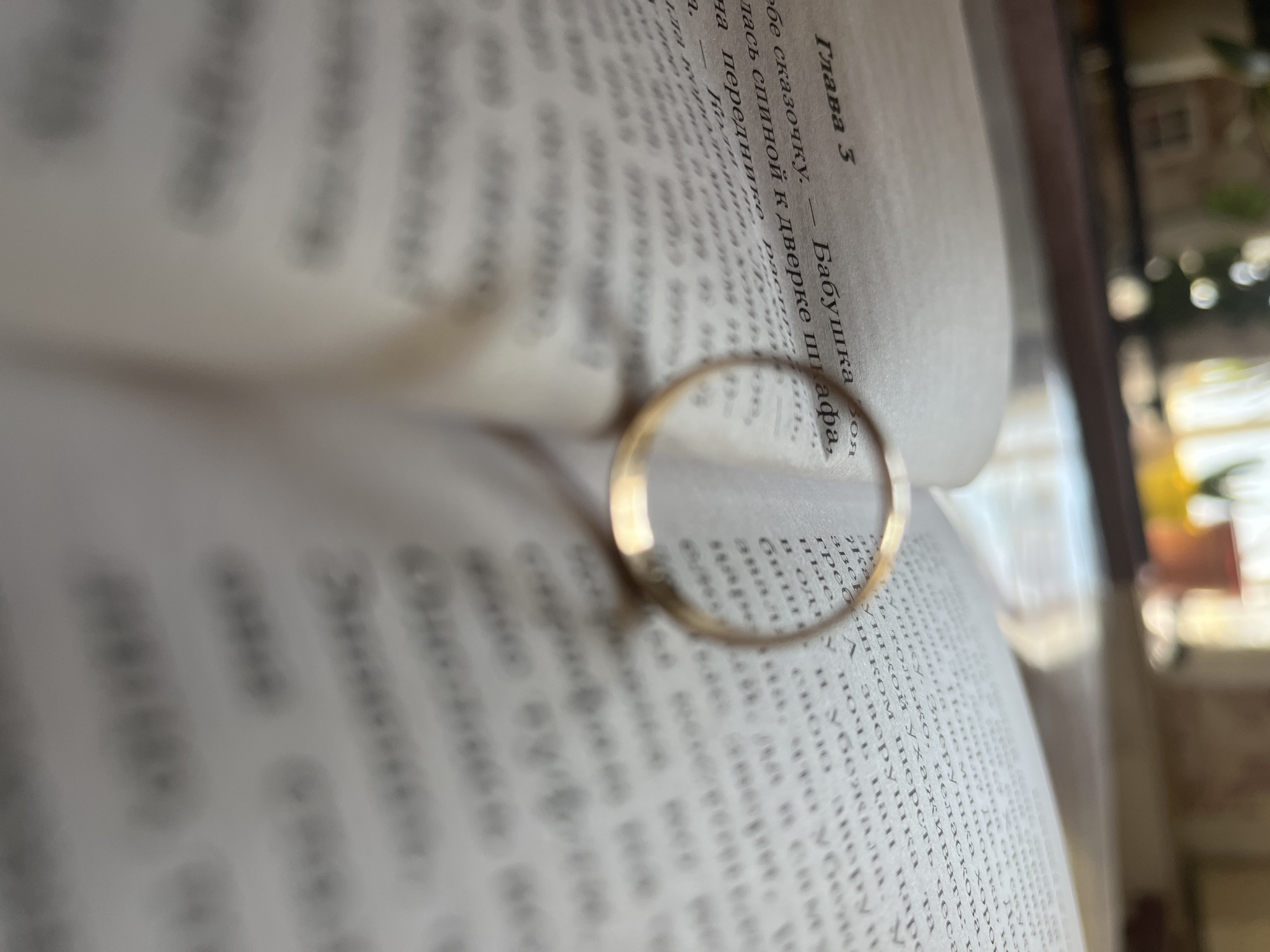 A close-up, shallow depth of field photograph shows an open book with Russian text. A gold-toned ring rests on the pages of the book, positioned horizontally across the center of the frame. The book appears to be lying on a surface, possibly a table, with a blurred background of indoor elements including light coming through a window, suggesting a domestic setting. The specific location of Krasnyye Barrikady, Russia, is not discernible from the image.