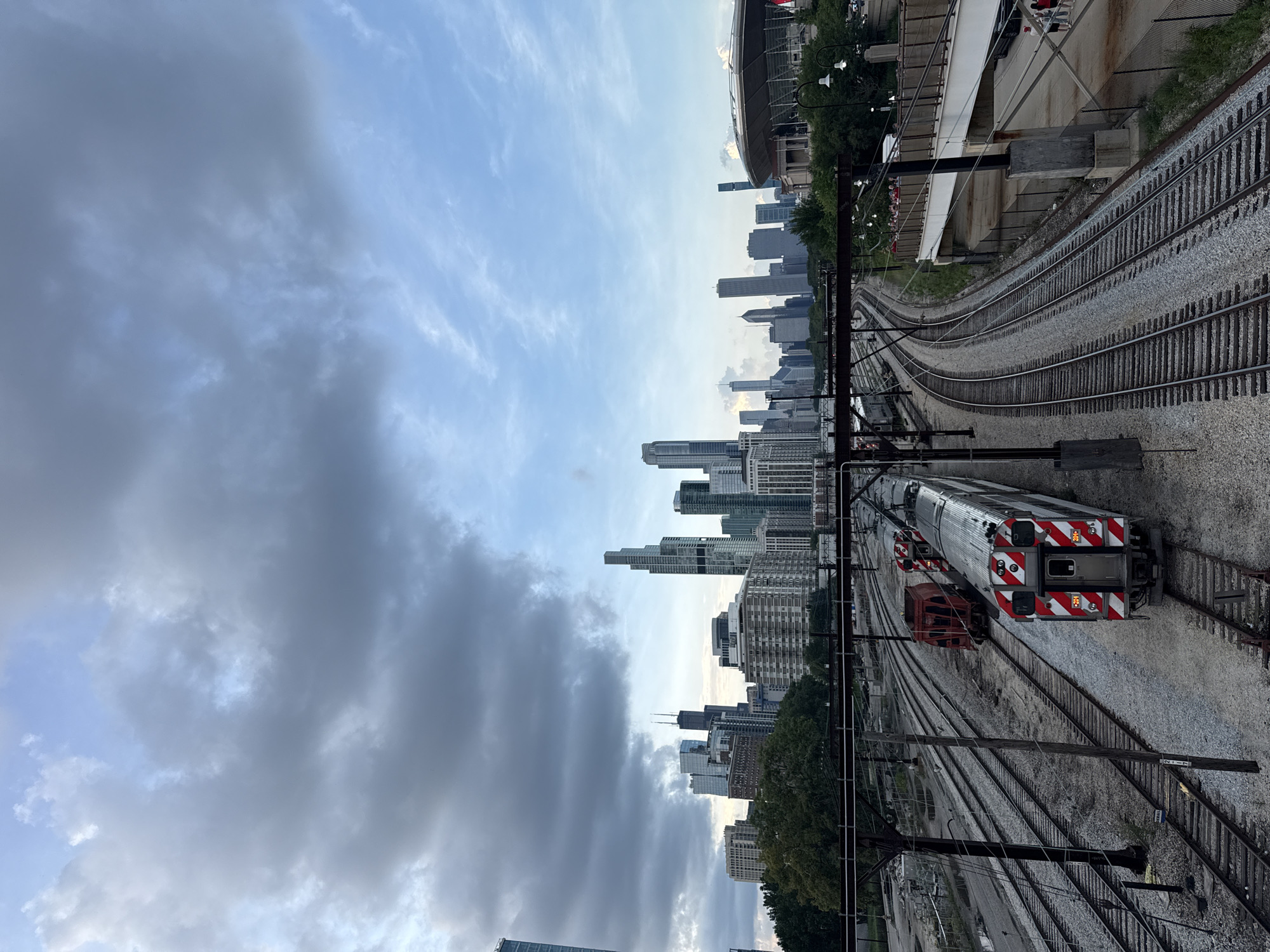 This image shows a view of the Chicago skyline with trains on railway tracks in the foreground. The skyline is dominated by tall buildings, including what appears to be Willis Tower and John Hancock Center. The trains are on multiple tracks, with one passenger train clearly visible, distinguished by its red and white striped livery. There are also some freight cars visible. The sky is partly cloudy with a mix of grey and blue hues. The scene is viewed from an elevated position, likely a bridge or overpass, looking towards the city. In the right side of the image, a stadium with a distinctive white roof is visible. The environment suggests an urban setting with significant infrastructure for transportation.