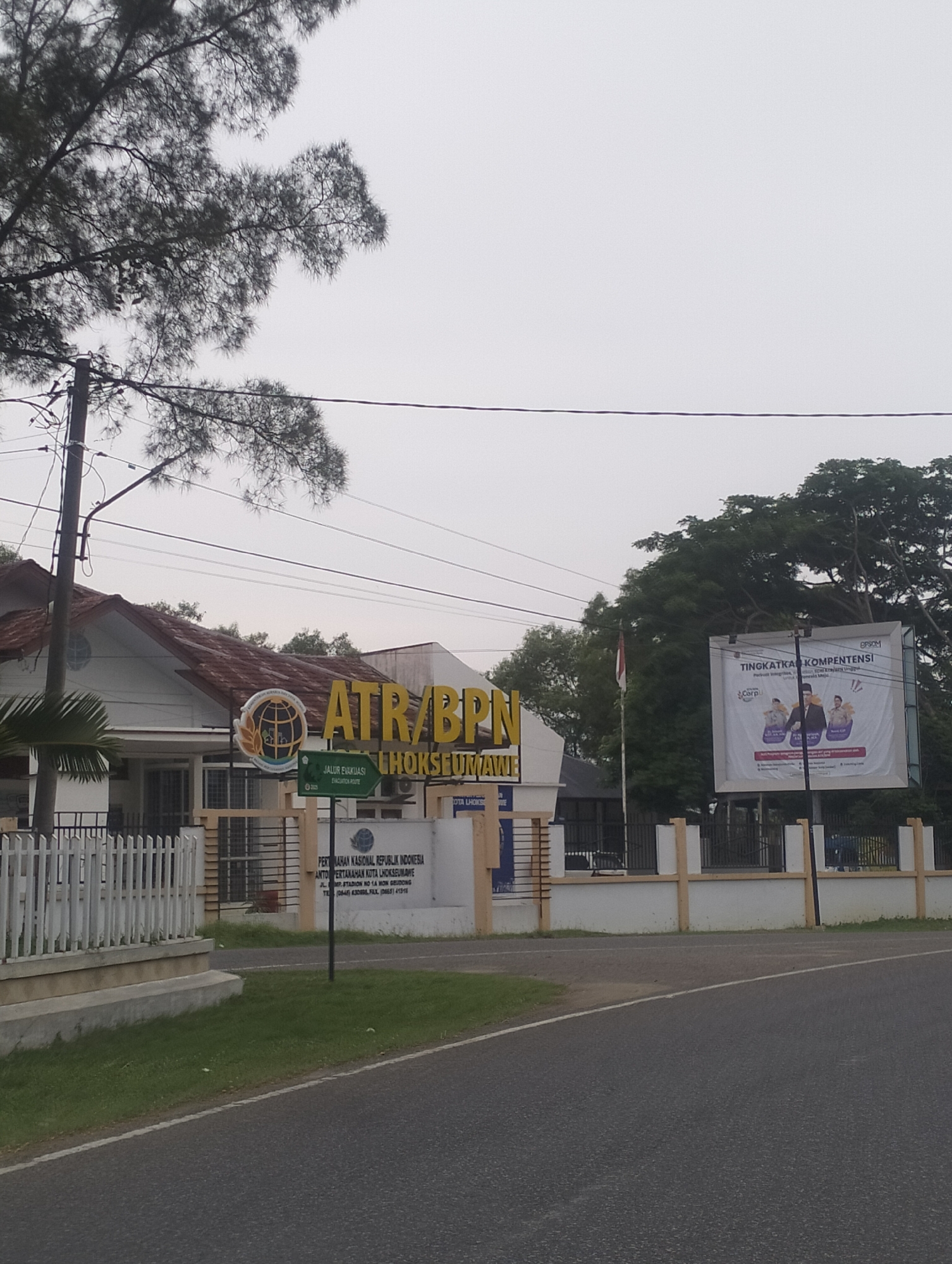 This is an outdoor, eye-level, full-shot photograph of the ATR/BPN office in Lhokseumawe, Indonesia. The building is white with a red-tiled roof. A sign above the entrance reads "ATR/BPN LHOKSEUMAWE". A white fence runs along the front of the property, and there is a green lawn in front of the fence. To the right of the building is a large billboard with text and images on it. The sky is overcast, and there are trees and power lines visible in the background. There are no people visible in the image.