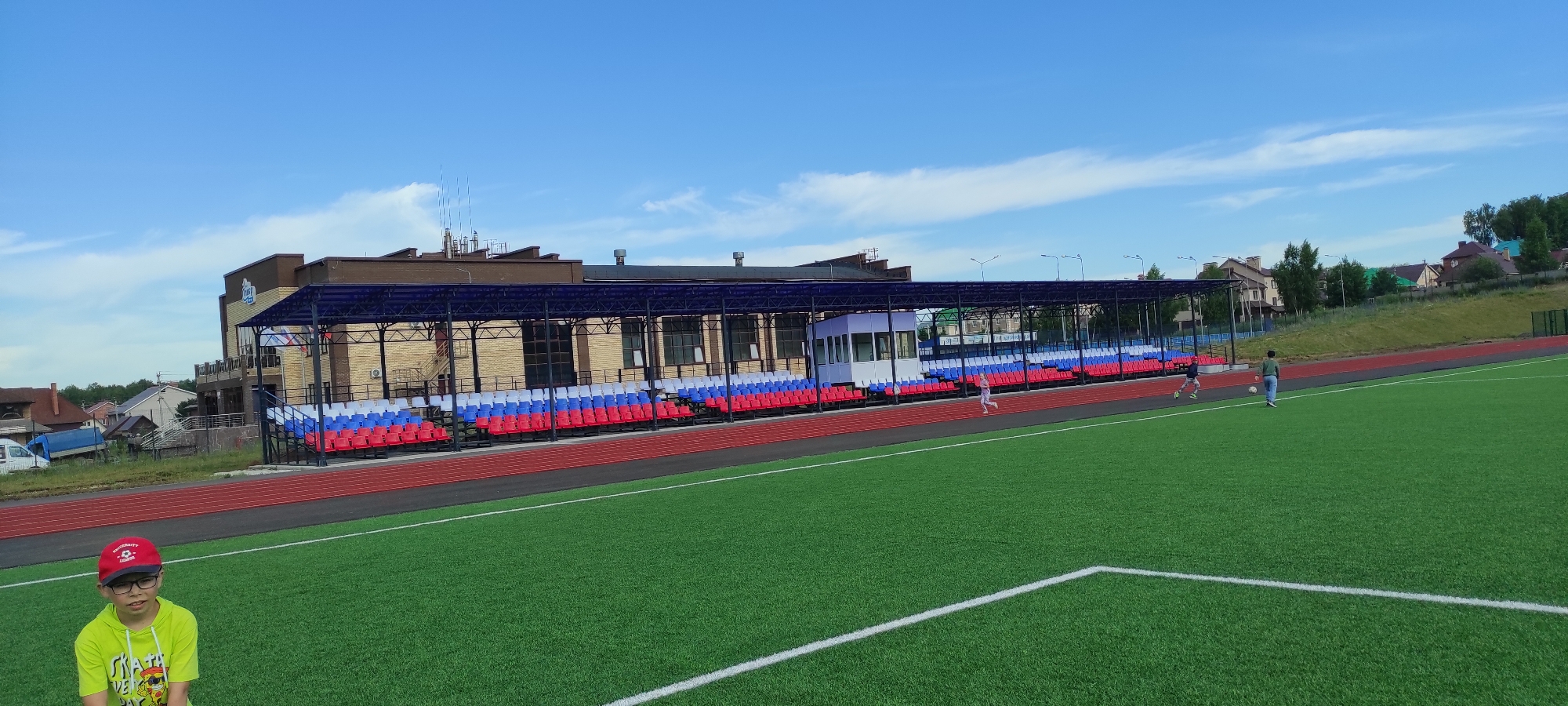 This is a full shot image of a sports stadium located in Noviy, Russia. The stadium features a green artificial turf field with white markings, a red running track, and tiered spectator seating. The seating is arranged in sections with red, white, and blue chairs, suggesting it may be the Russian national colors. A covered grandstand with a blue roof structure extends along one side of the field. A small white booth or press box is visible beneath the grandstand.
In the foreground, a young boy wearing glasses and a red baseball cap with a logo stands facing the camera, dressed in a bright yellow hooded sweatshirt.
In the middle ground and background, several people are visible on the running track and the field. Two children appear to be running on the track near the grandstand, and an adult is seen kicking a soccer ball on the field with another child running towards them.
The stadium is bordered by a grassy embankment on the right side and a residential area with houses and trees in the distance on the left. The sky is blue with scattered white clouds. The overall impression is that of a modern, well-maintained sports facility.