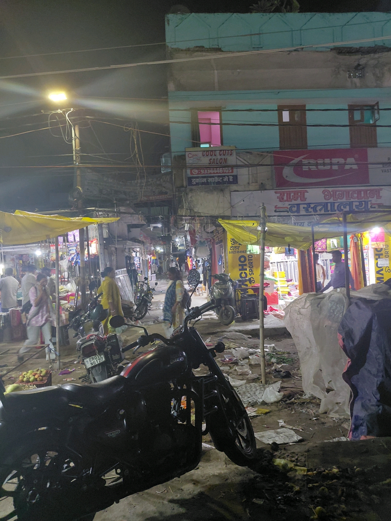 The image depicts a street scene at night in Patna, India, possibly in a market area. A bright street lamp illuminates the foreground and midground. In the foreground, a black motorcycle with red accents is parked, dominating the lower half of the frame. Its details, such as the "Royal Enfield" branding on the fuel tank and the "Classic" badge on the side, are visible. Behind the motorcycle, another motorcycle with a visible license plate "BR 01 N6127" is parked.
The midground features a bustling street with several stalls covered by yellow tarps. People are visible, some walking and some seemingly engaged in transactions. One person in a yellow tunic is prominent, standing near a stall. Another person in a pink and white checkered shirt is walking towards the left.
The background shows a multi-story building with light blue and white facade. Signs in Hindi are visible on the building, including "RAM BHAGWAN VASTRALAYA" and "COOL CUTS SALON." The windows of the building are illuminated, one with a pink interior light. The street itself appears cluttered with debris, including plastic bags and other refuse, suggesting an active but potentially unkempt environment. The overall lighting suggests it is nighttime, with artificial light sources dominating the scene.