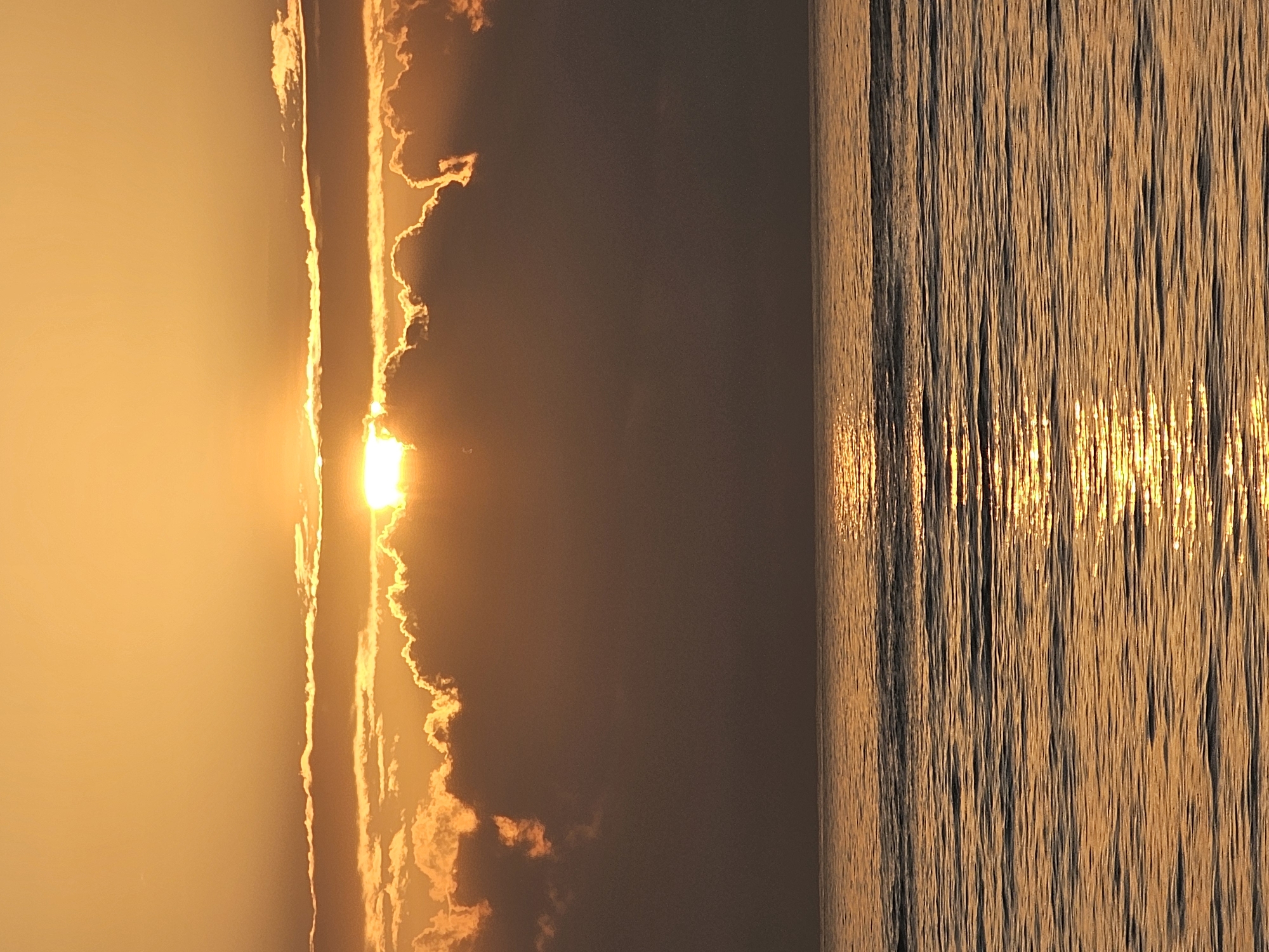 The image depicts a sunset over the ocean, likely near Denpasar, Indonesia, given the atmospheric conditions and common coastal scenery. The sun is partially obscured by thick clouds, casting a warm, golden light across the sky and reflecting vibrantly on the rippling water. The horizon line is clearly defined, separating the turbulent cloudscape from the serene ocean surface. The water itself is a deep, rich orange, with sunlight creating a shimmering path across its waves. There are no people, subjects, objects, or actions visible in the scene, making it a purely environmental capture. The overall impression is one of natural beauty and tranquility at the close of day.
