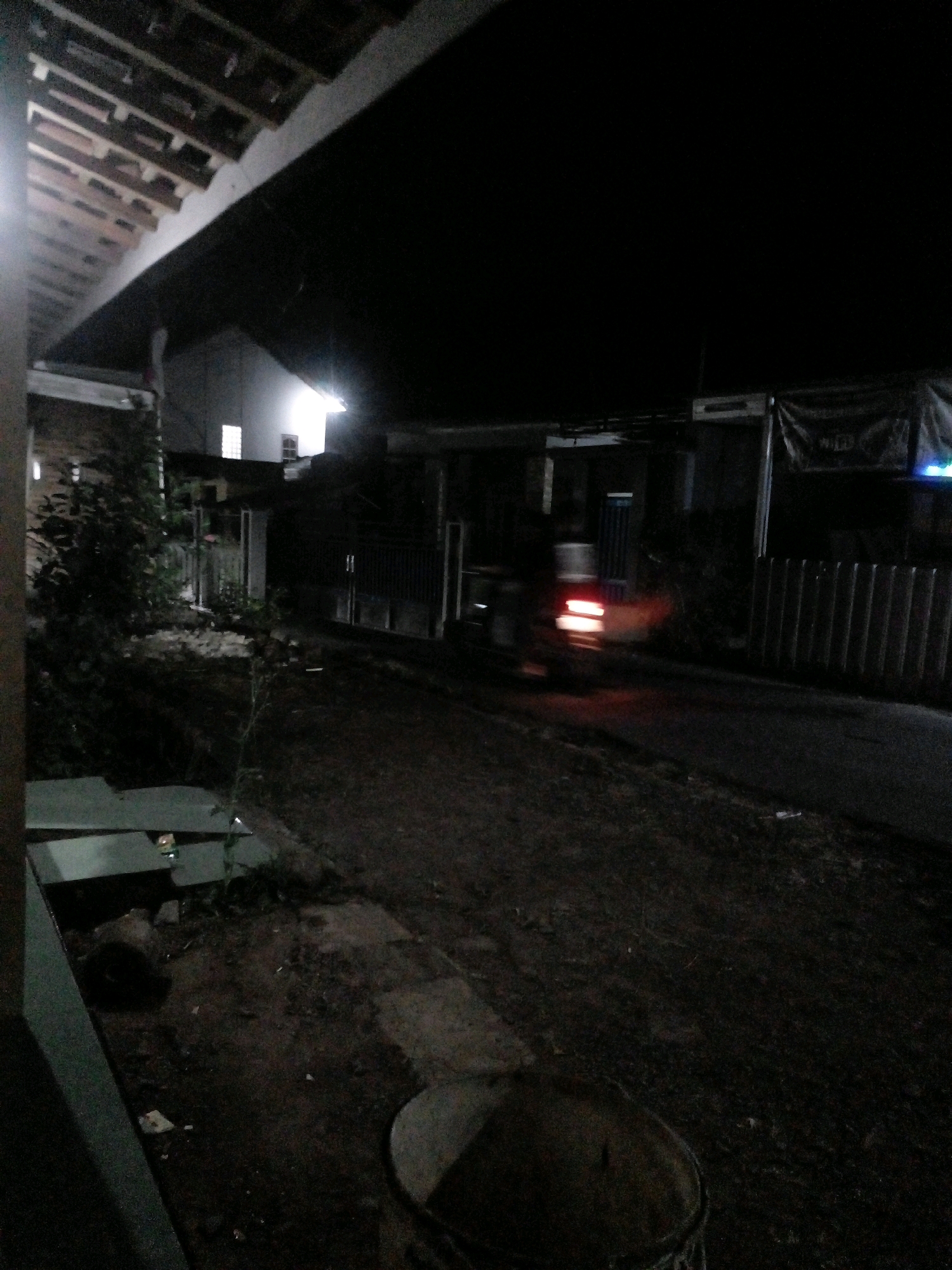 The image captures a nighttime scene on a street in Pungangan, Indonesia. A vehicle, blurred due to motion, is visible on the asphalt road, its red taillights emitting a glow. To the left of the road, there is a dark, uneven ground with some debris and foliage. A light-colored, rectangular structure, possibly a bench, is present in the foreground left. Beyond the road, houses with illuminated windows can be seen. The architecture suggests a residential area. A black banner with white text is hanging on a building on the right side of the street, partially obscured by darkness. The overall atmosphere is dark and quiet, indicative of a residential neighborhood at night.