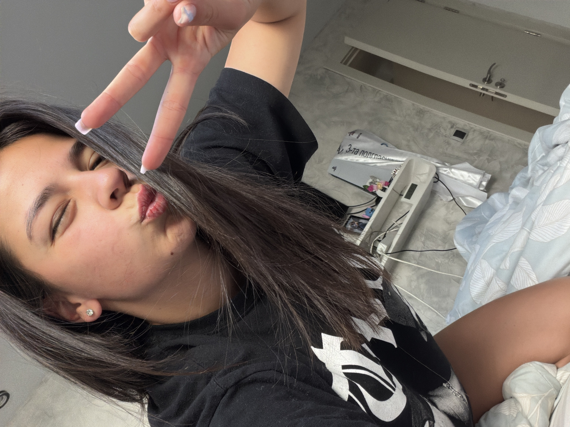 A woman with dark hair is holding up a peace sign while pursing her lips for a selfie. She is wearing a black t-shirt with a white design. She has fair skin and a small earring in her left ear. The background includes a bed with light blue and white bedding, a white shelf unit, and a grey wall with a white fixture. There is a box on the shelf with text in Cyrillic script that includes the words "за подрасна" suggesting the location is in a country that uses the Cyrillic alphabet, but Gabrovo, Bulgaria cannot be discerned from the image.